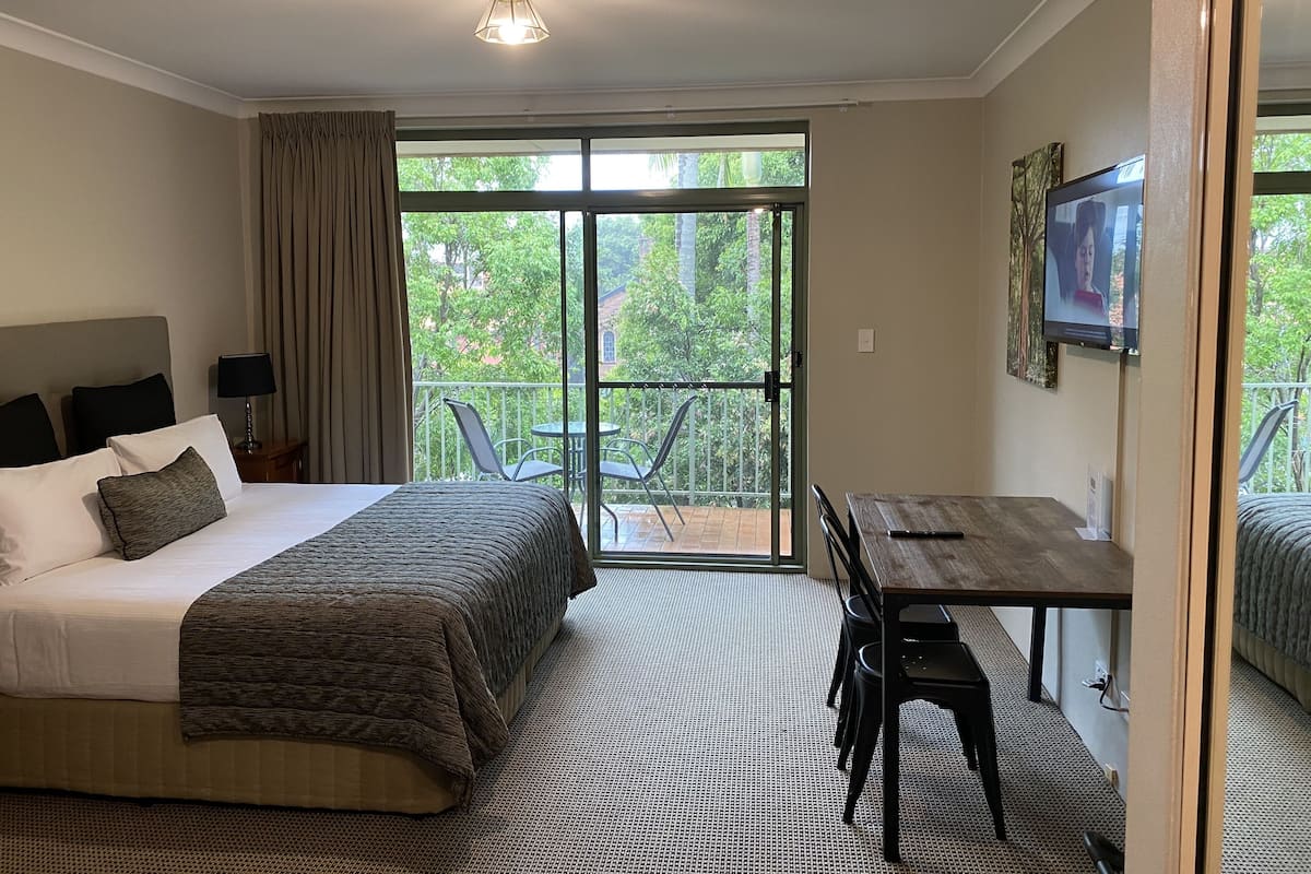 Habitación de hotel con cama, escritorio, televisor y balcón con vista a árboles.