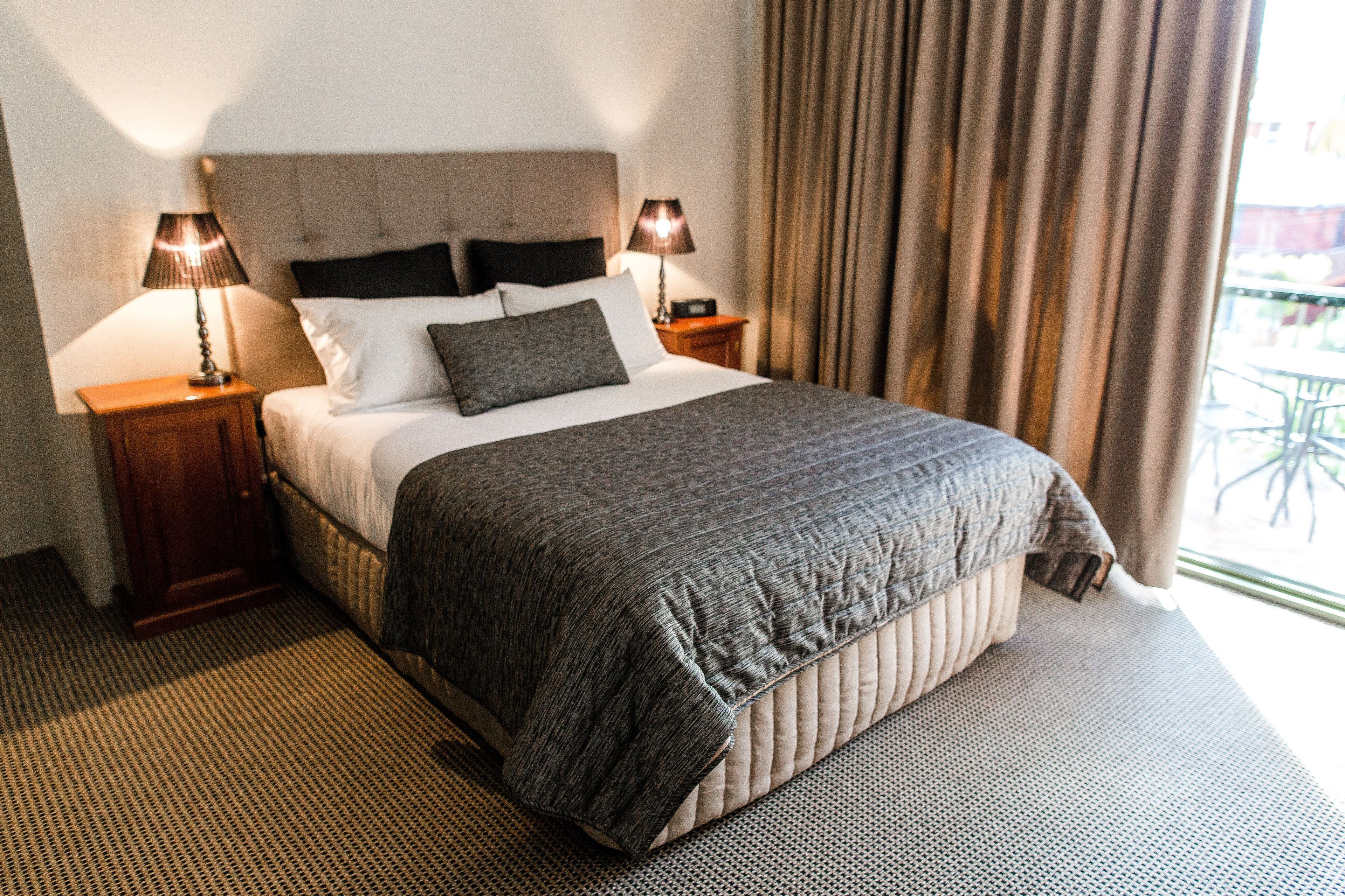 A hotel room with a large bed, two bedside tables with lamps, a headboard, and a view of the outdoors through curtains.