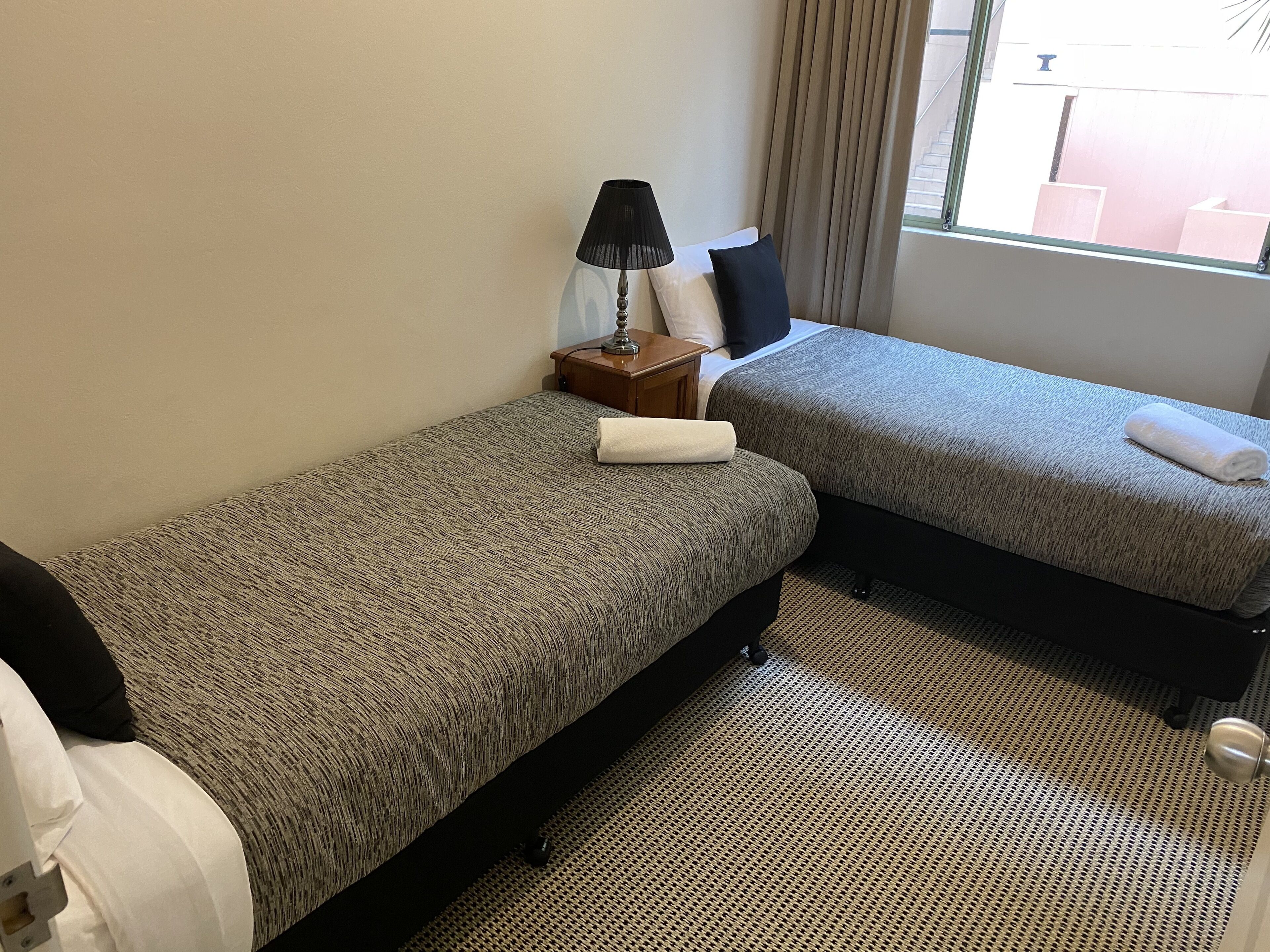 A hotel room with two single beds, a bedside table with a lamp, a window with curtains, and a view of the outside.