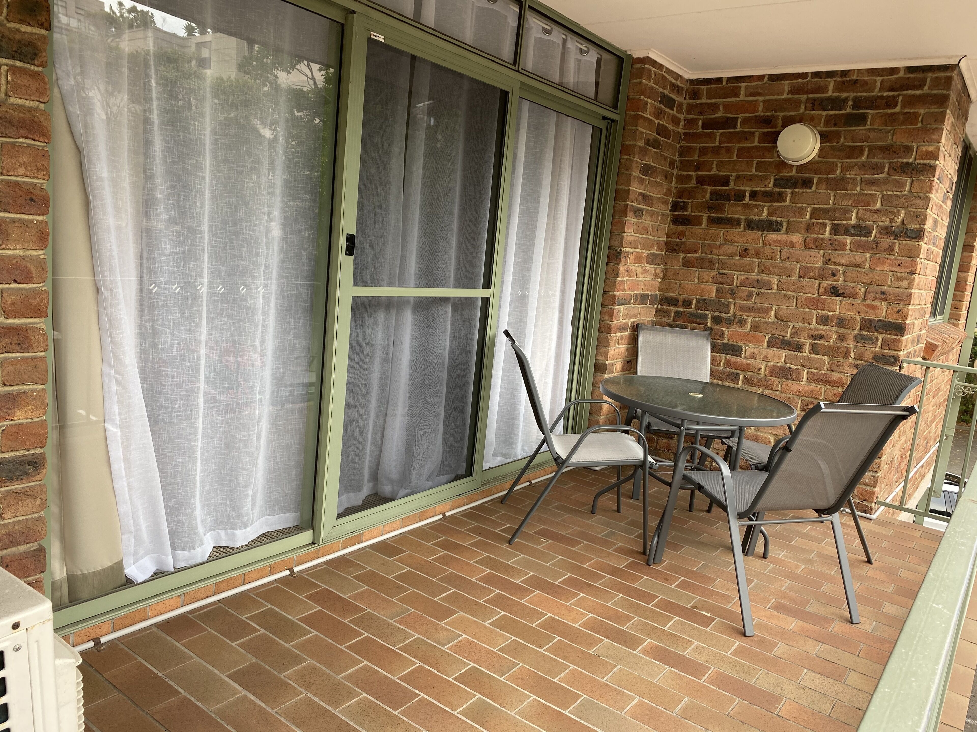 A balcony with a brick wall, a sliding glass door with white curtains, and a round table with four chairs.