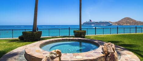 A swimming pool with a stone border, surrounded by grass and palm trees, with a view of the ocean and a cruise ship in the distance.
