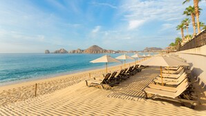 Een strand met rijen ligbedden en parasols, palmbomen en een helder uitzicht op de oceaan en verre bergen.