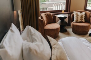 A hotel room with a bed, two armchairs, a small table with a glass, and a view of the outside. - Hotel Omega Amsterdam (Amsterdam)