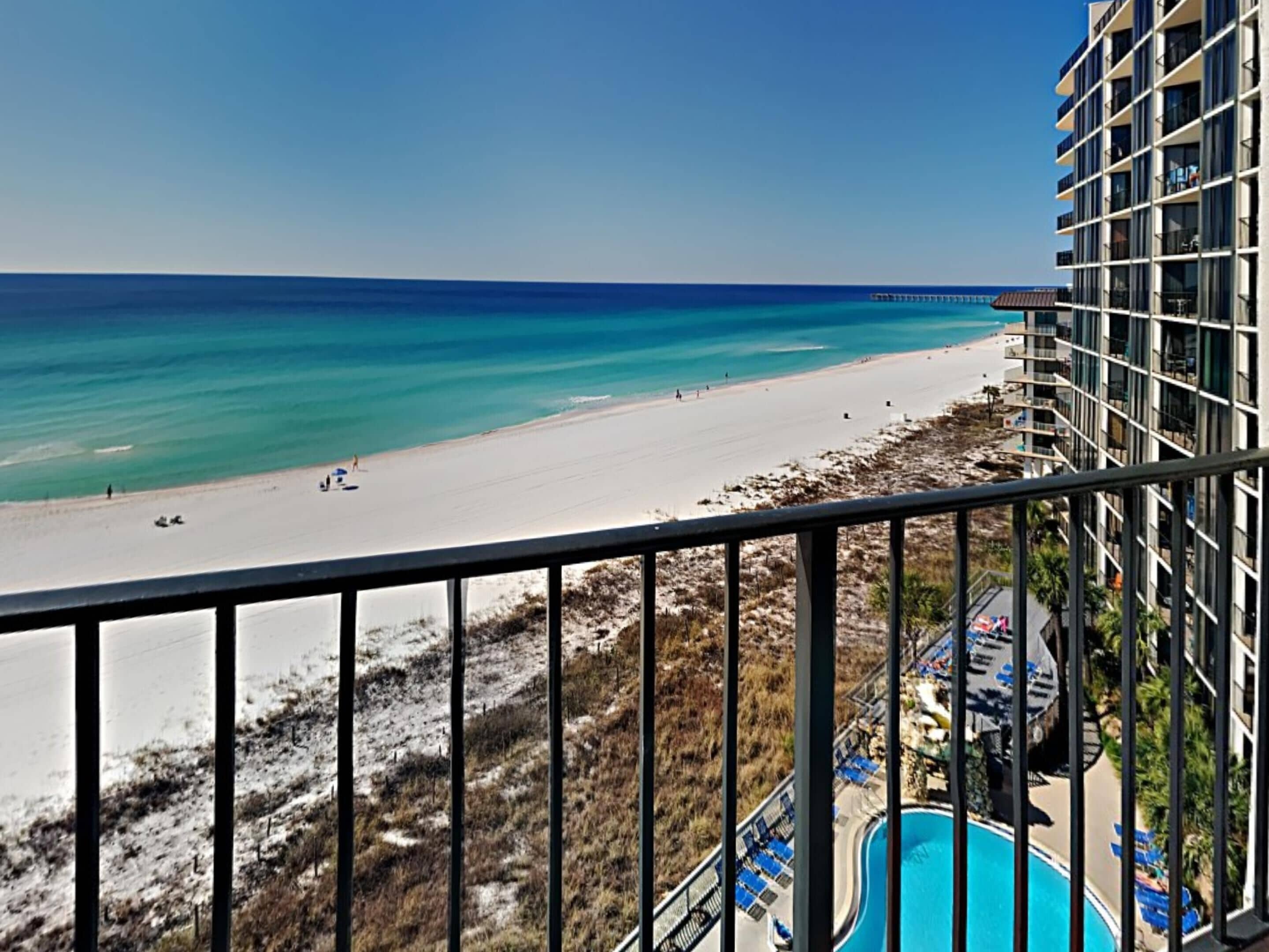 A beach view from a balcony overlooking a swimming pool and a sandy beach with a clear blue ocean.