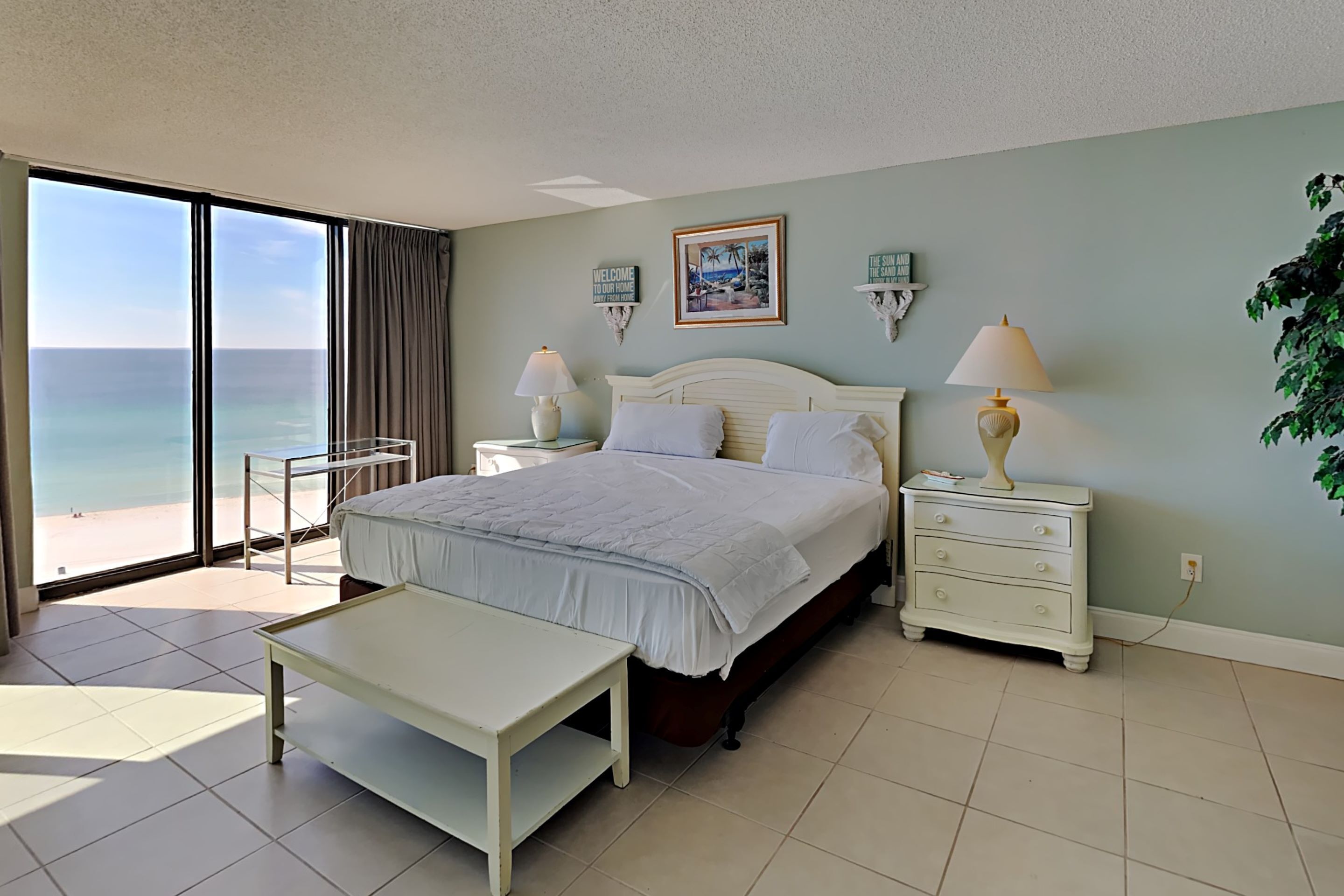 A bedroom with a bed, bedside tables, a nightstand, a bench, and a view of the ocean.