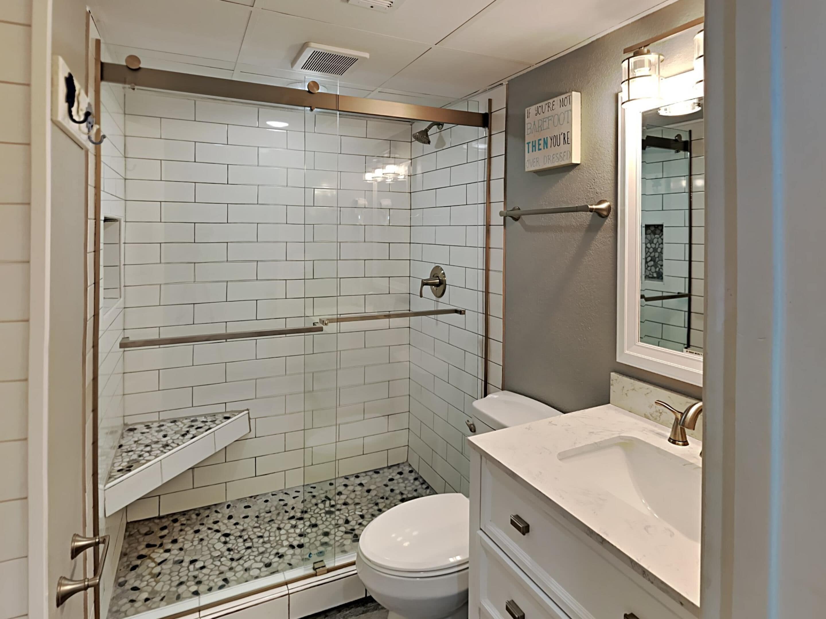 A bathroom with white subway tiles, a glass shower enclosure, a toilet, a sink with a marble countertop, and a mirror with a light above it.