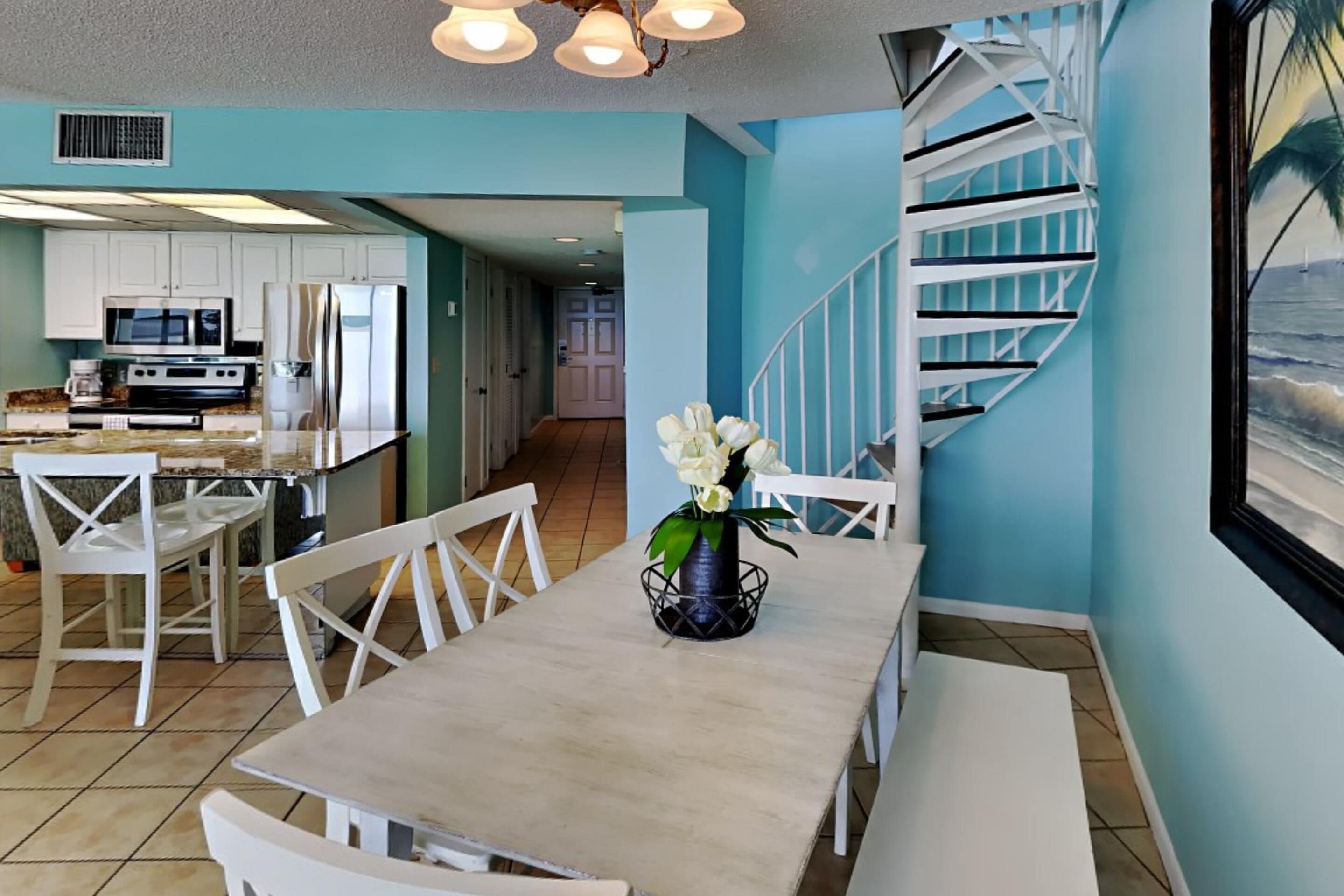 A dining area with a table, chairs, and a vase of flowers. There is a kitchen in the background with white cabinets and appliances.