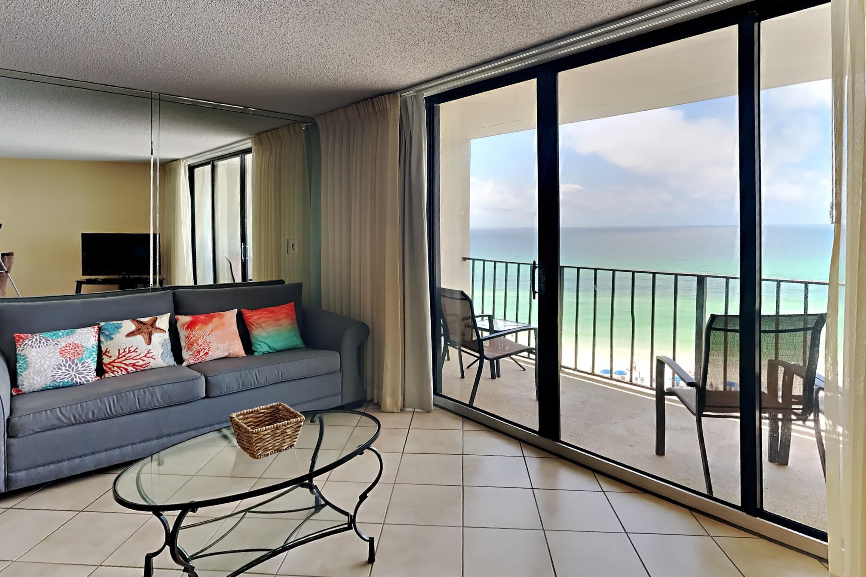 A hotel room with a balcony, a sofa with colorful cushions, a glass coffee table, and a TV.