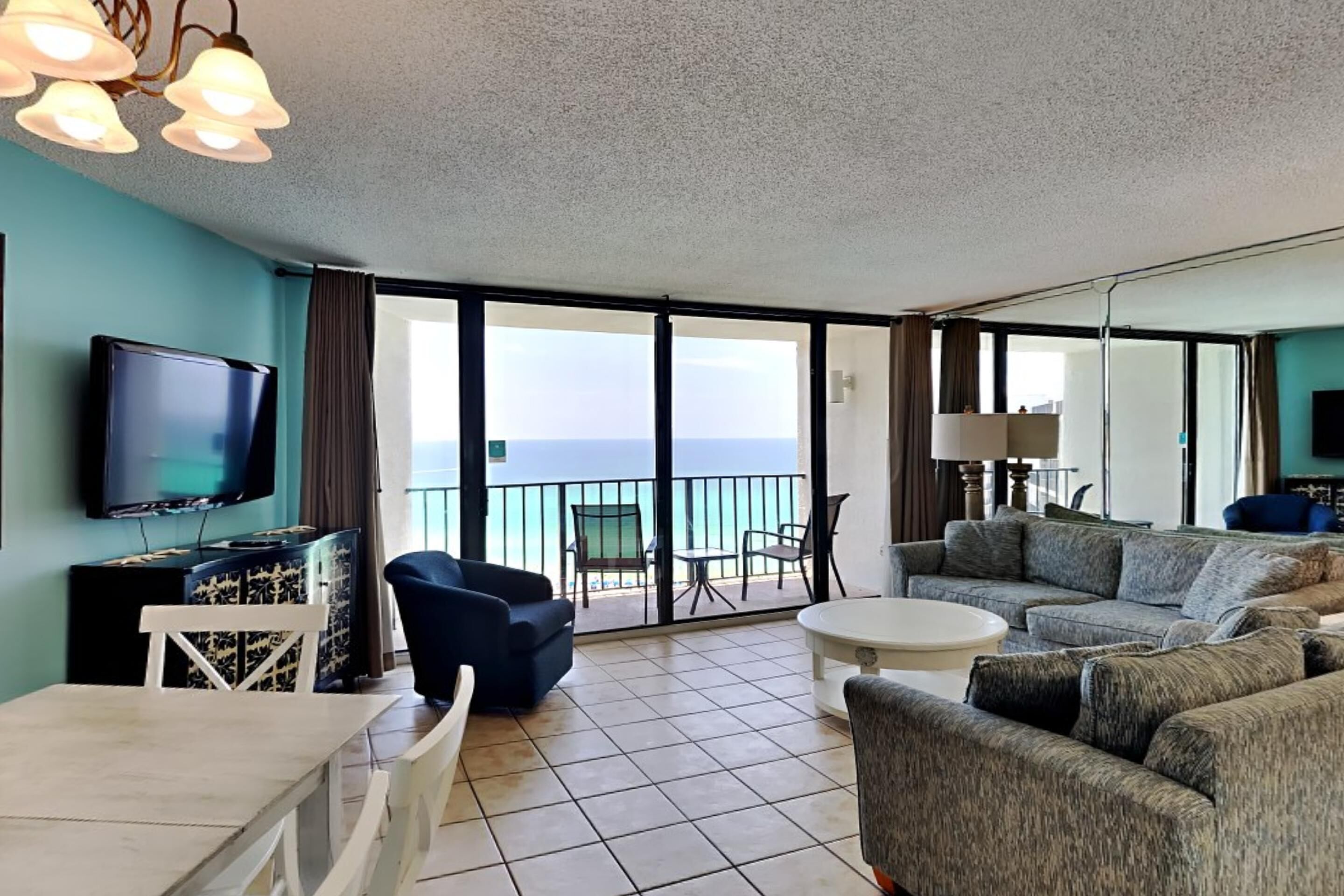 A spacious living area with a large flat-screen TV, a dining table with chairs, a sofa, and a balcony with a view of the ocean.