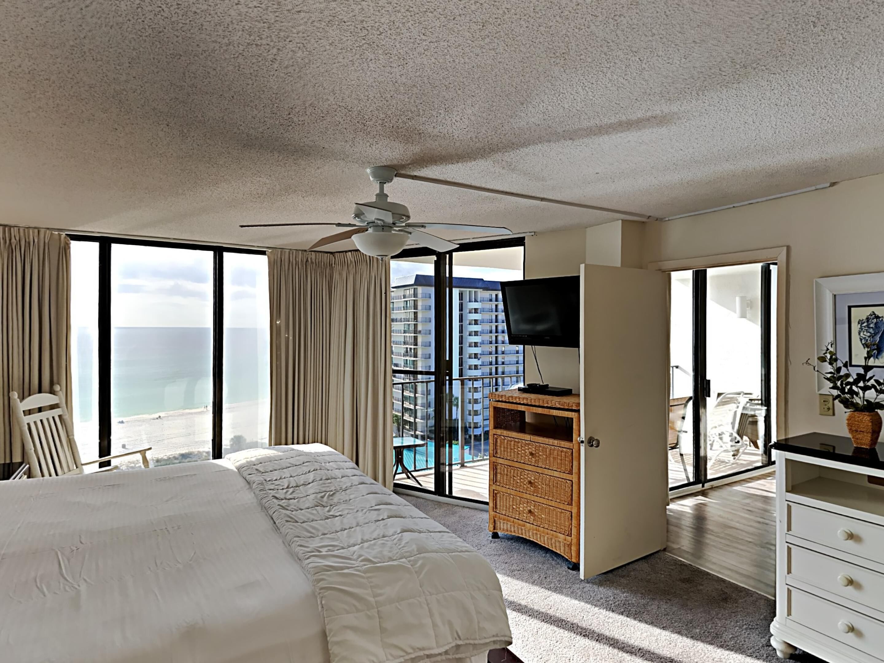A hotel room with a bed, a ceiling fan, a TV, a dresser, and a view of the ocean and buildings.