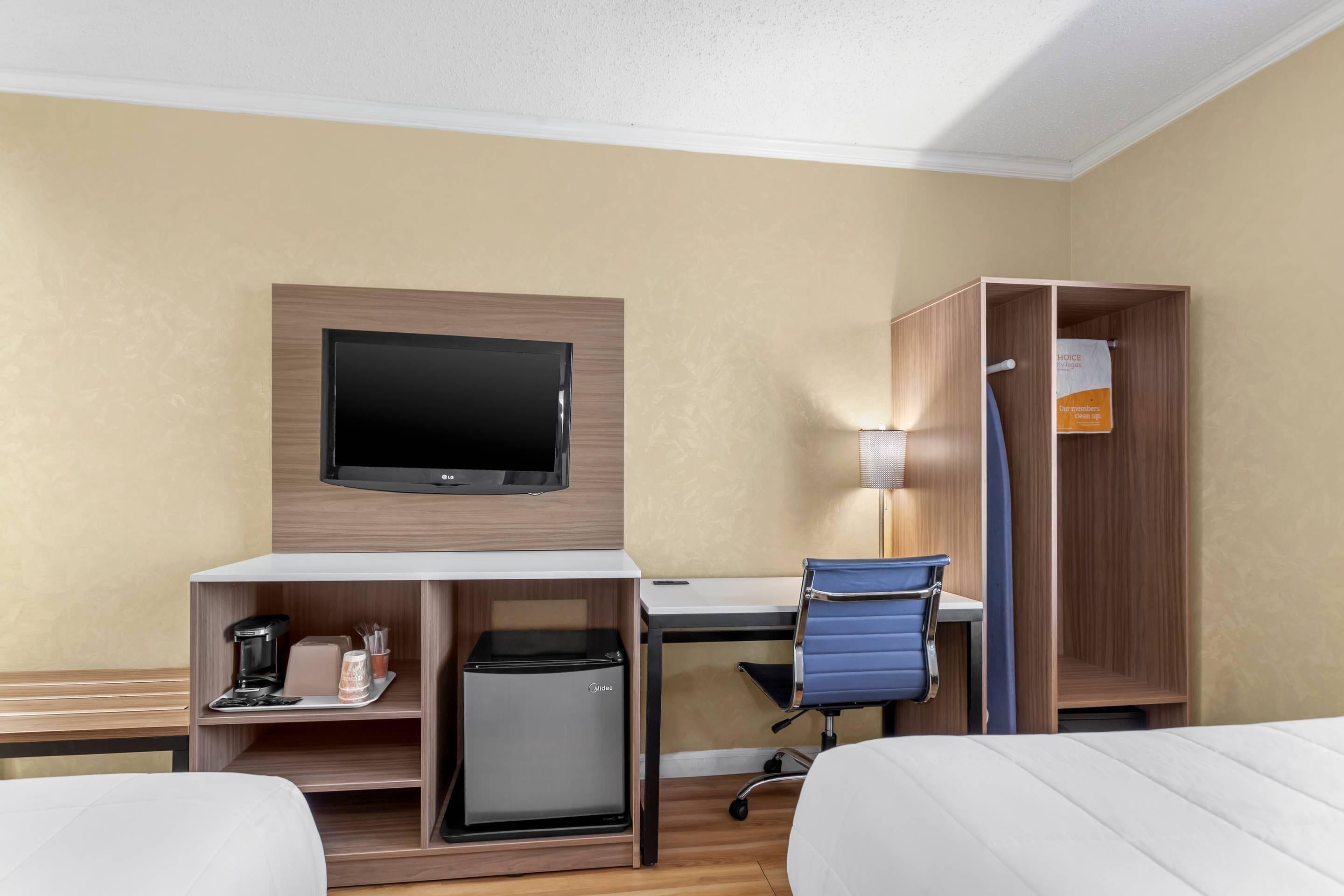 Habitación de hotel con cama, escritorio con computadora, televisor, mini refrigerador, cafetera y armario.
