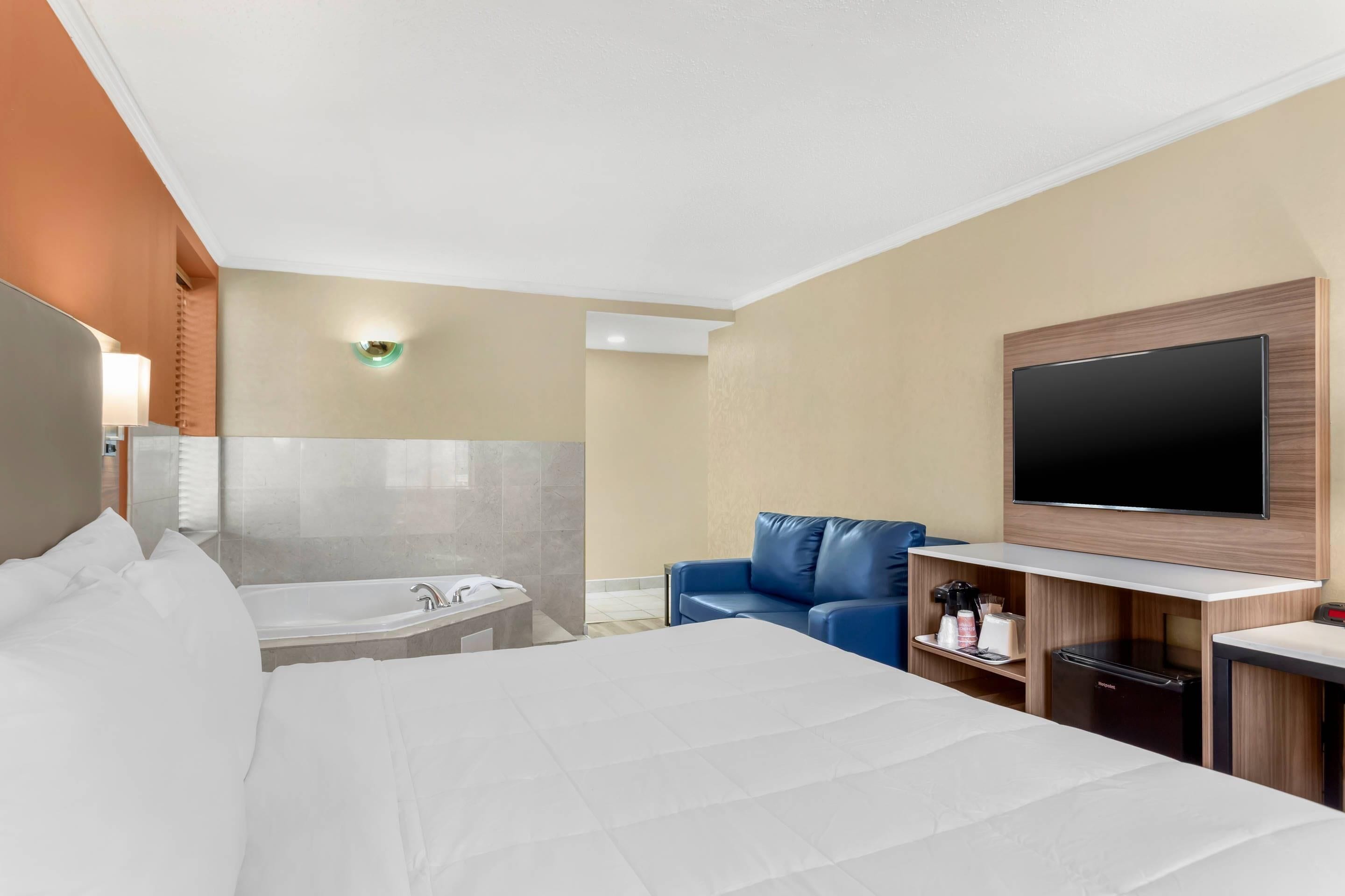 A hotel room with a bed, a desk, a television, a sofa, a bathtub, and a minibar.