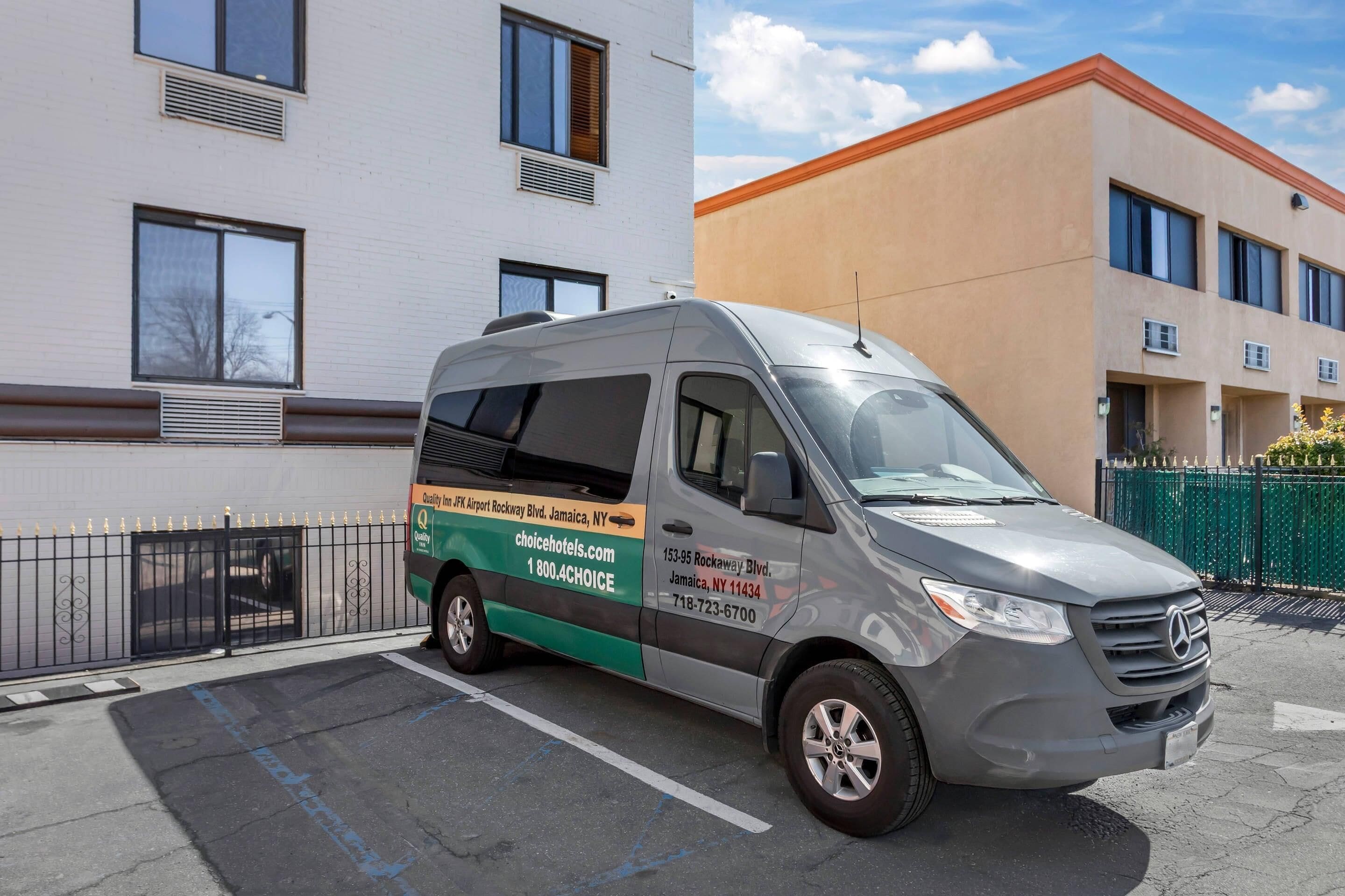 A gray van parked in front of a building with a white and beige facade, a black metal fence, and a clear sky.