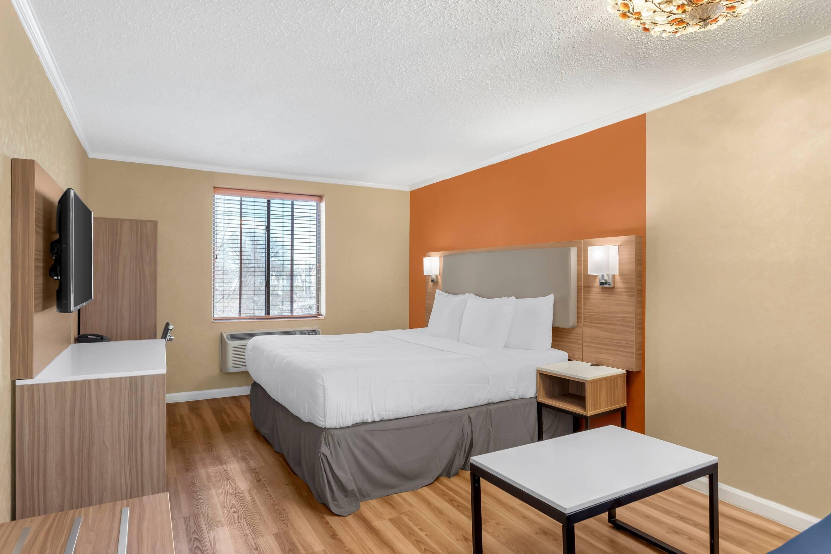 A modern hotel room with a bed, bedside tables, a TV, a window with blinds, and a ceiling light fixture.