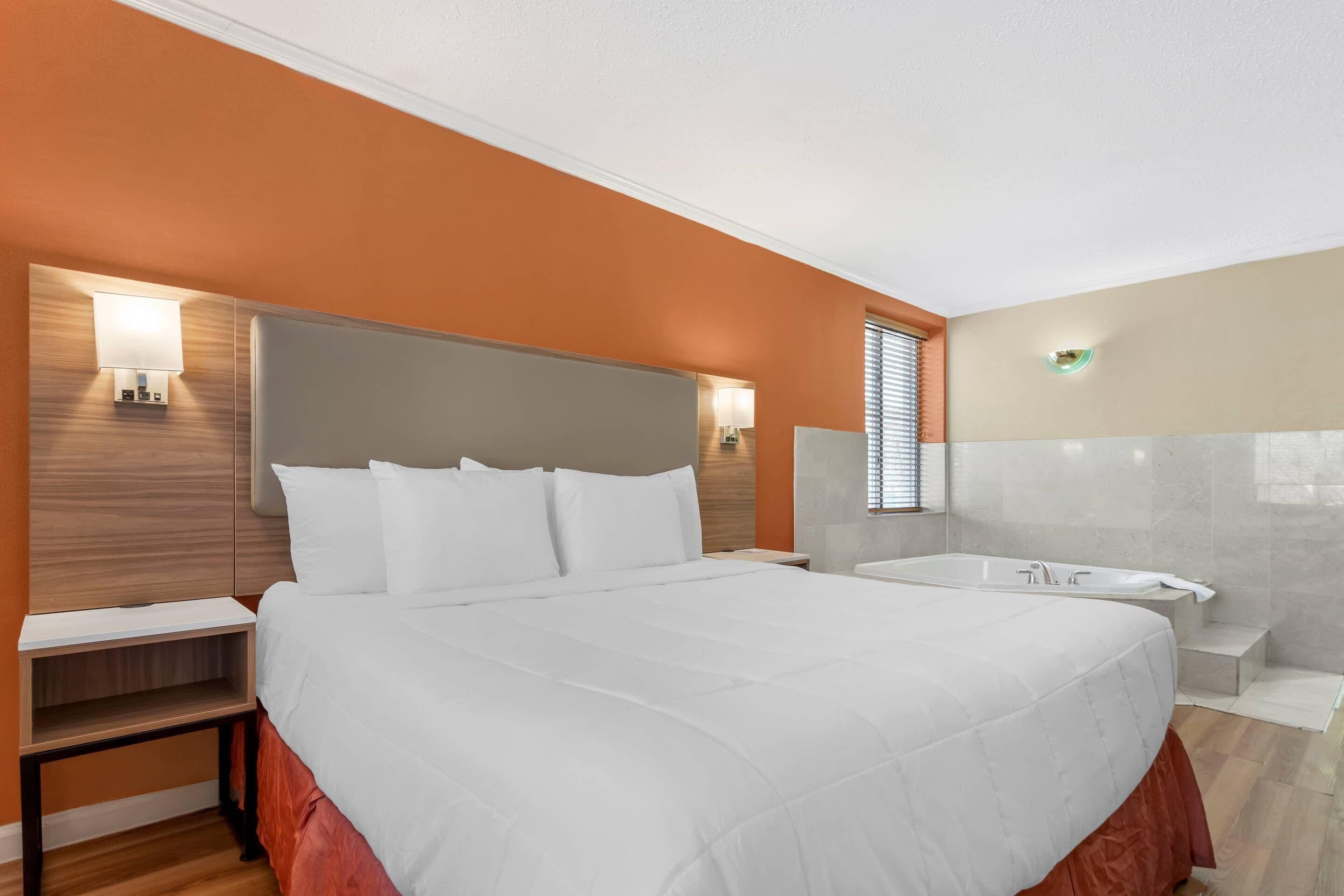 A modern hotel room with a large bed, a built-in desk, a vanity with a mirror, and a bathtub.