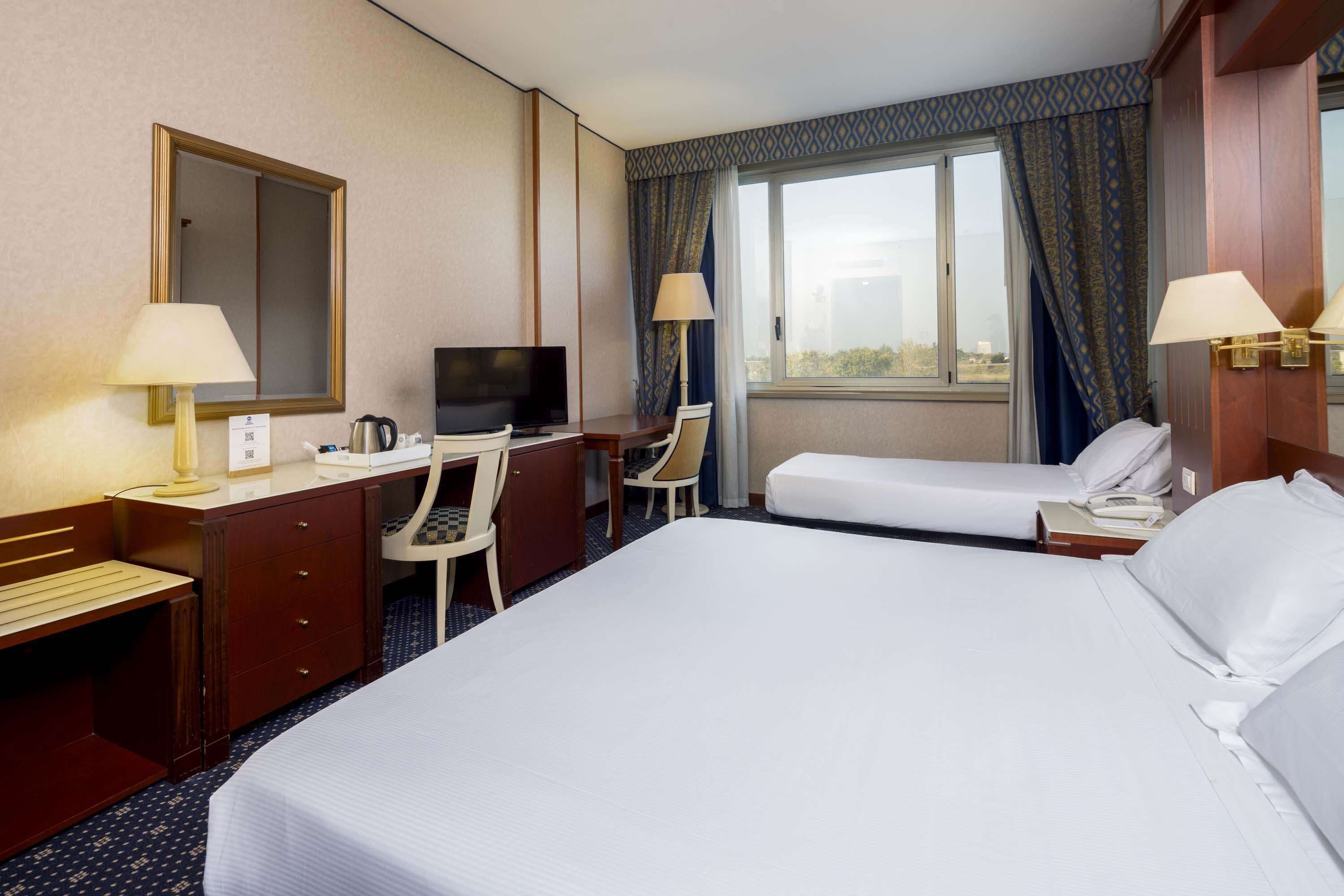 A hotel room with a large bed, a desk with a TV, a chair, a desk lamp, and a window with curtains.