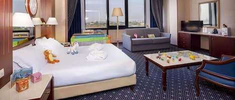 A hotel room with a bed, a sofa, a desk with a TV, and a table with children's toys.