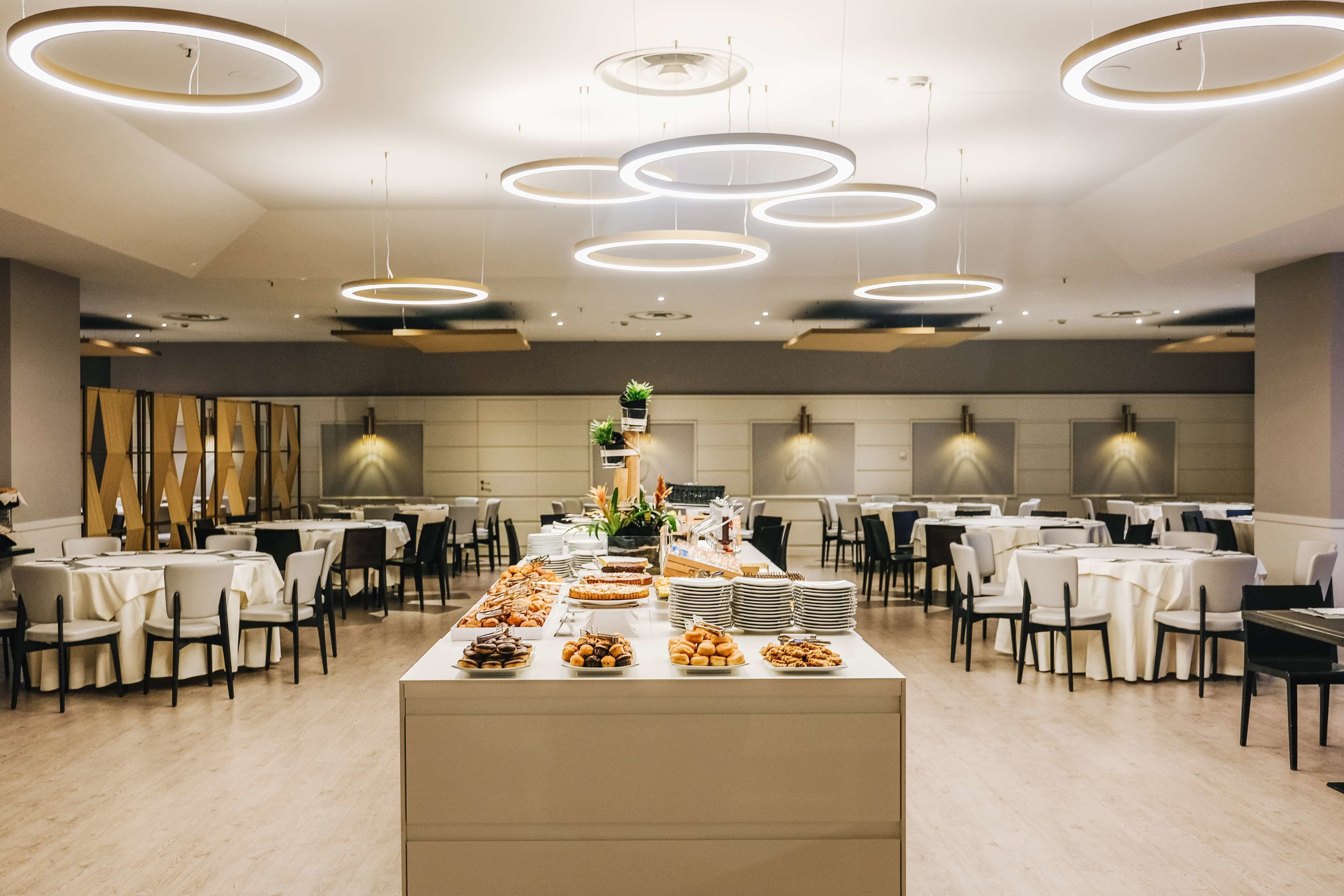 A spacious banquet hall with multiple round tables and chairs, a central buffet table with various food items, and modern circular ceiling lights.