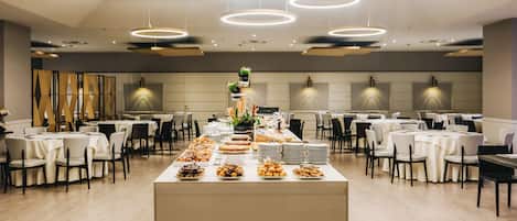 Un amplio salón de banquetes con varias mesas redondas y sillas, una mesa de buffet central con diferentes opciones de comida y modernas lámparas circulares en el techo.