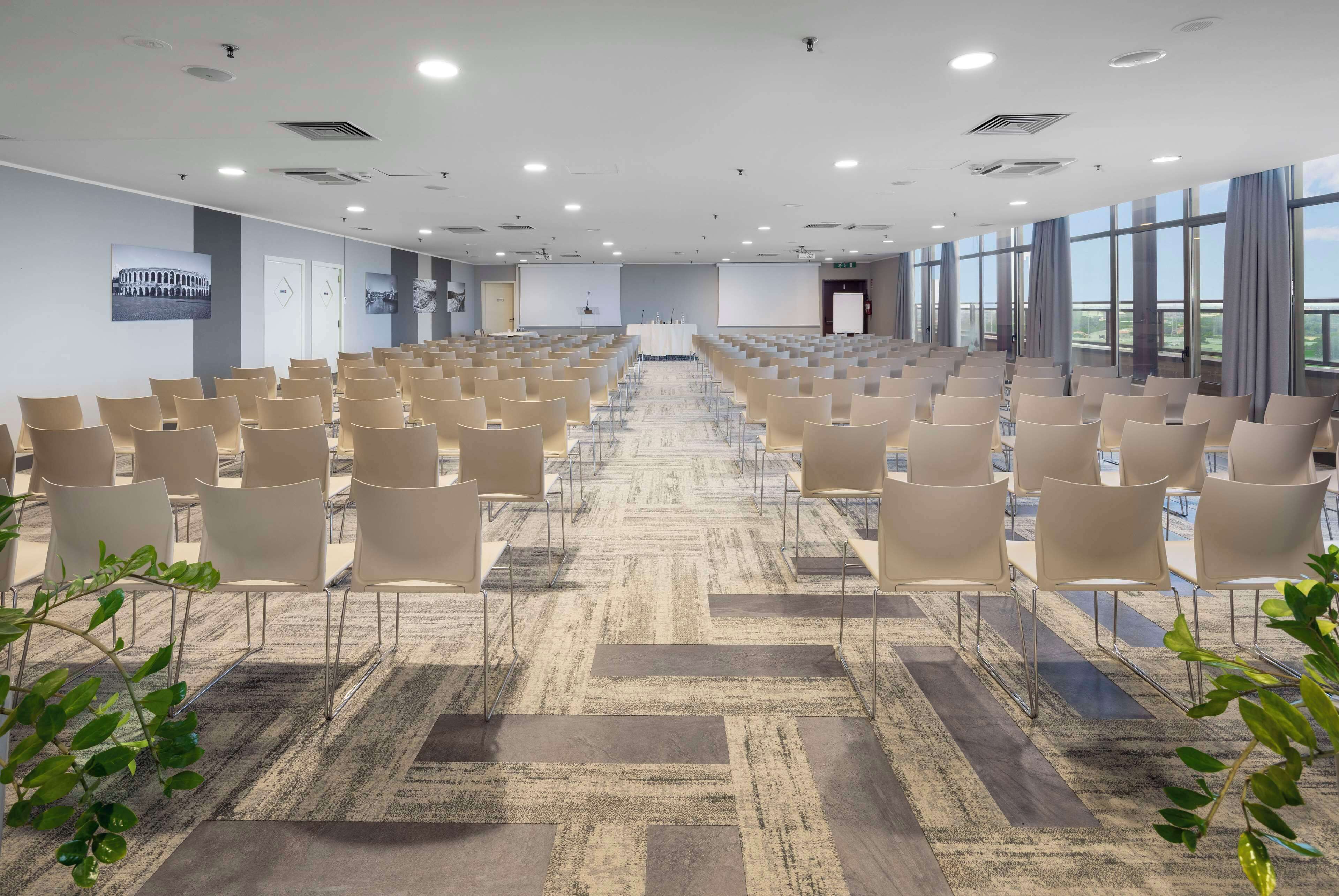 Uma sala de conferências com fileiras de cadeiras, piso coberto de carpete e amplas janelas.