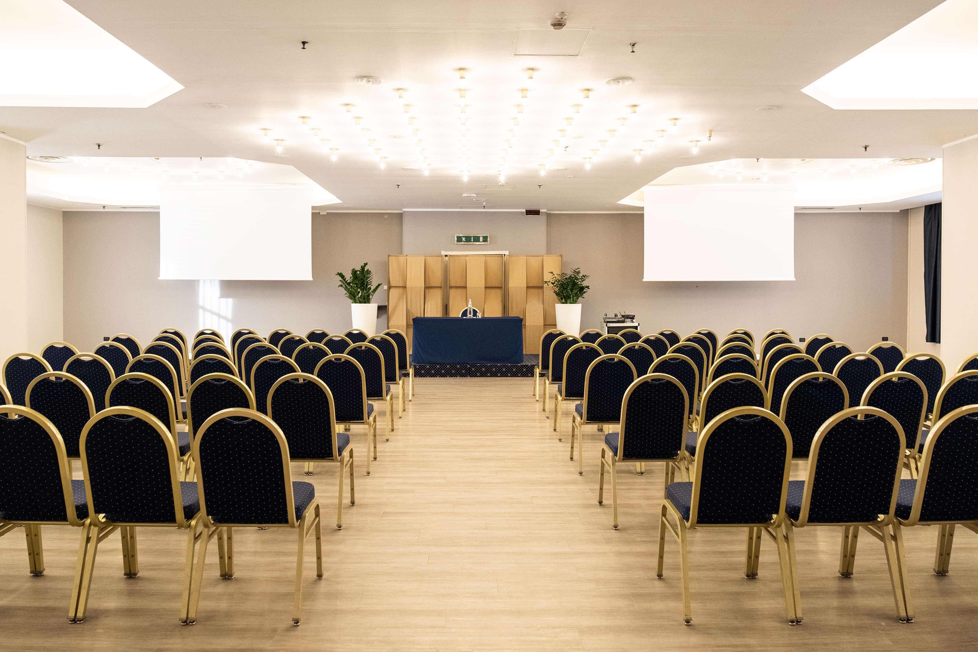 A conference room with rows of chairs facing a stage, a podium, and potted plants.