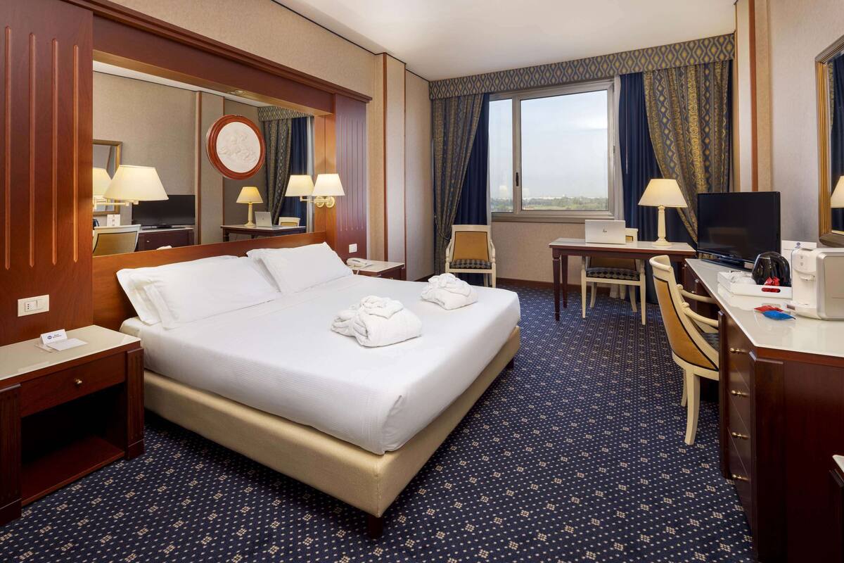 Ein Hotelzimmer mit einem großen Bett, einem Schreibtisch mit Computer, einem Sessel und Blick auf die Stadt durch das Fenster.
