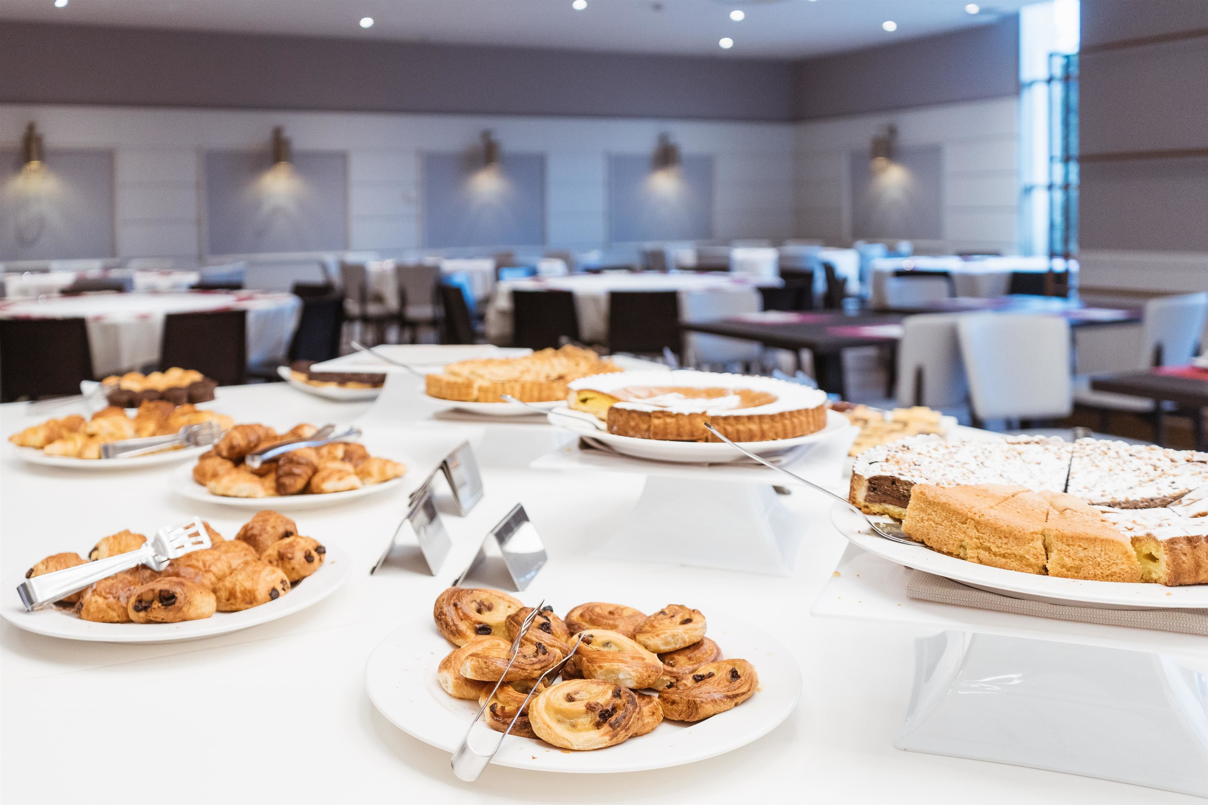 A buffet with various pastries and desserts, including croissants and a large pie, in a well-lit dining area with tables and chairs.