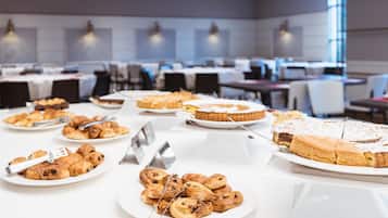 Un bufé con diversos pasteles y postres, incluyendo croissants y una tarta grande, en un comedor bien iluminado con mesas y sillas.