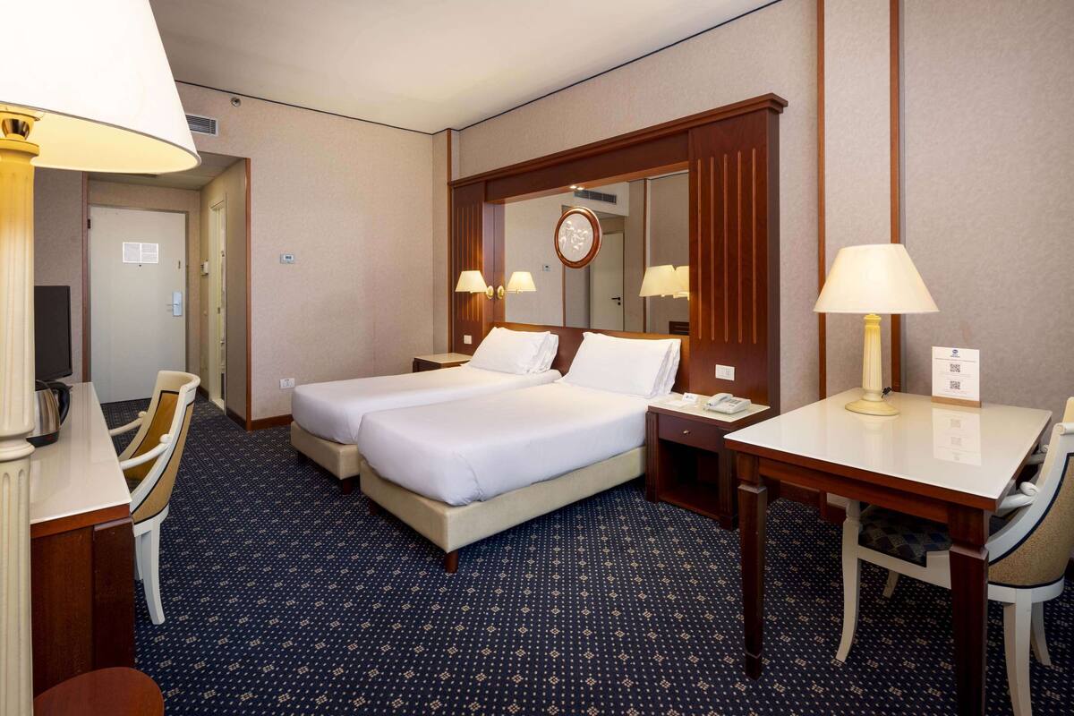 Ein Hotelzimmer mit zwei Betten, einem Schreibtisch, einem Stuhl, einer Lampe und einem Spiegel.