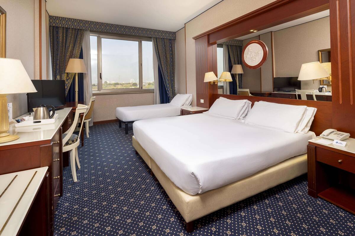 Ein Hotelzimmer mit einem großen Bett, zwei Nachttischen, einem Schreibtisch mit Stuhl, einem Fernseher und einem Fenster mit Vorhängen.