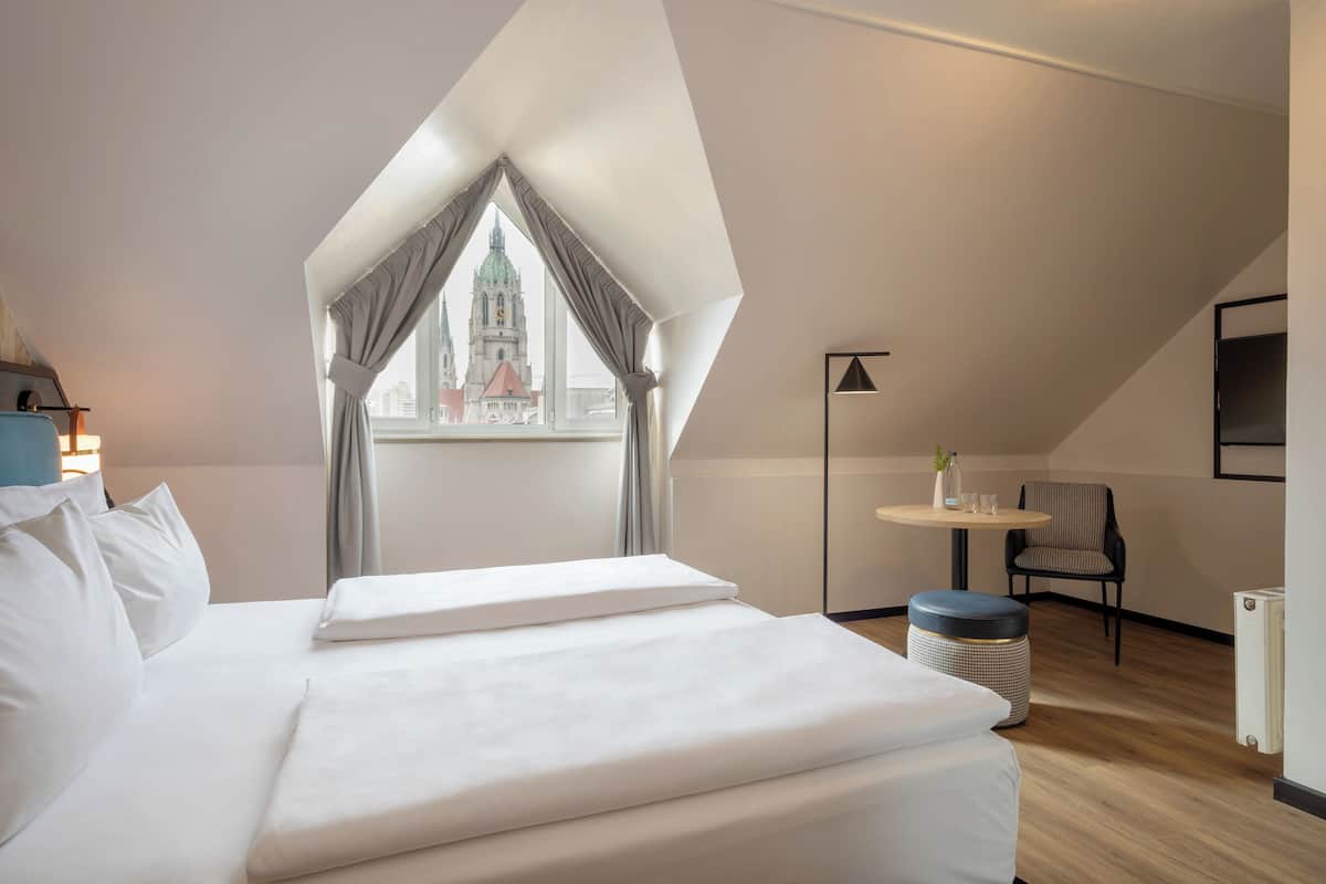A modern hotel room with a large bed, a small round table with a chair, a view of a historic building, and a skylight.
