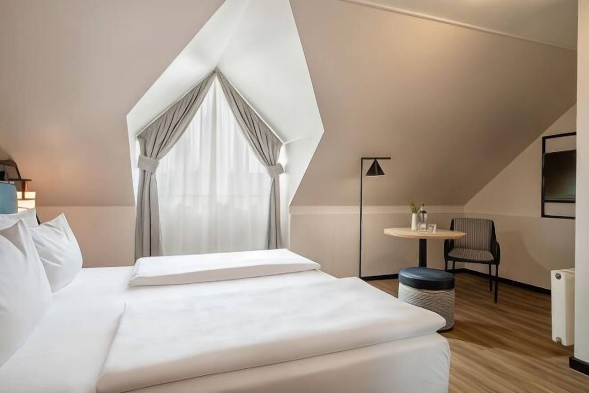 A modern hotel room with a large bed, a small round table, a chair, and a view of the outdoors.