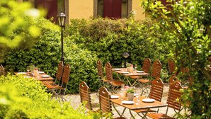 An outdoor dining area with tables and chairs, surrounded by greenery and a building in the background. - Hotel München City Center affiliated by Meliá (Munich)