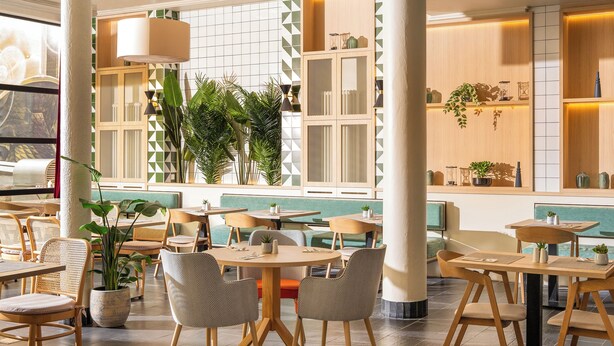 A modern dining area with round tables and chairs, potted plants, and a view of the sea through large windows. - Hotel München City Center affiliated by Meliá (Munich)