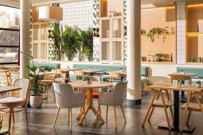 A modern dining area with round tables and chairs, potted plants, and a view of the sea through large windows. - Hotel München City Center affiliated by Meliá (Munich)