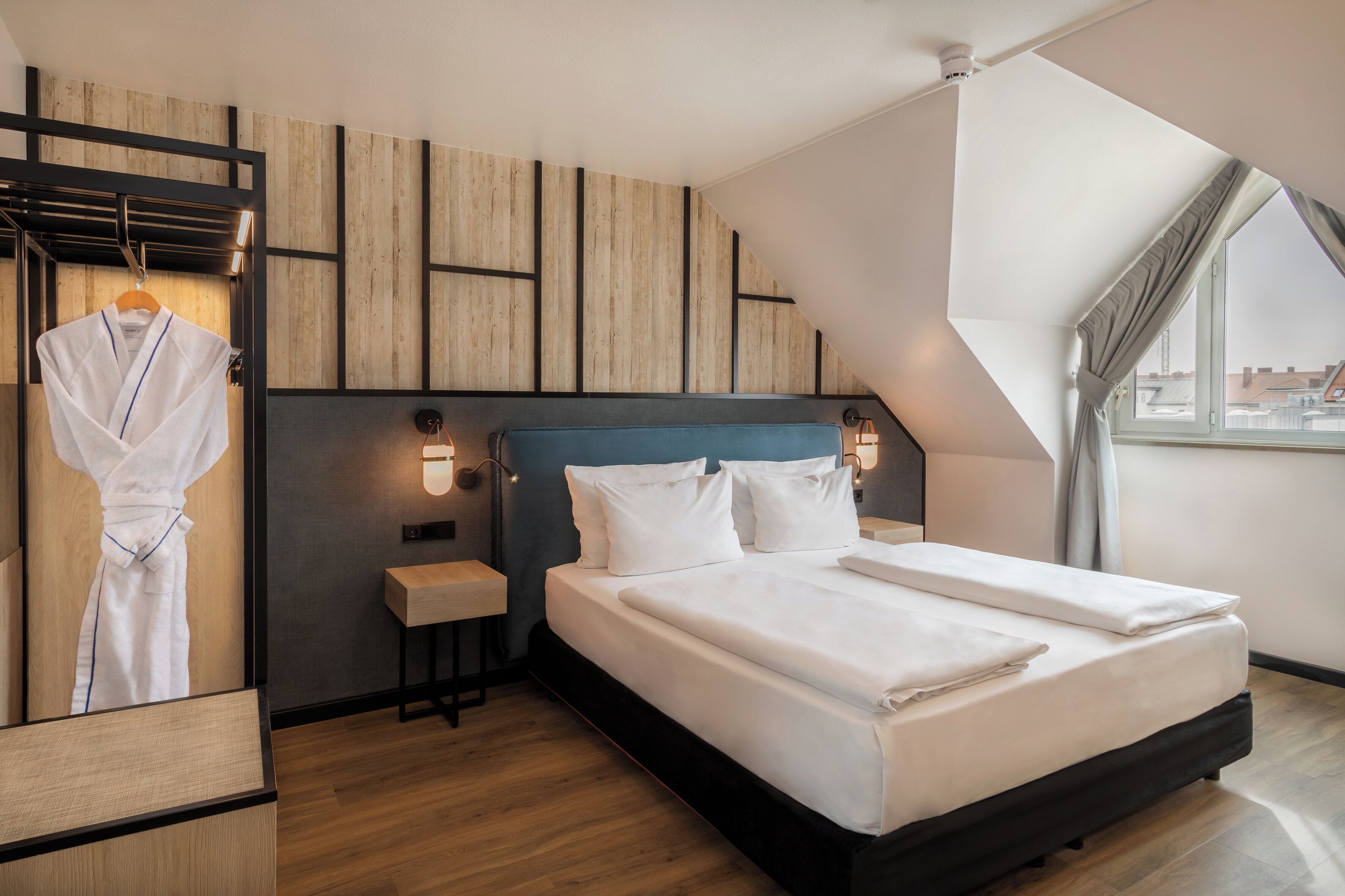 A modern hotel room with a large bed, a wooden wardrobe, and a small bedside table.