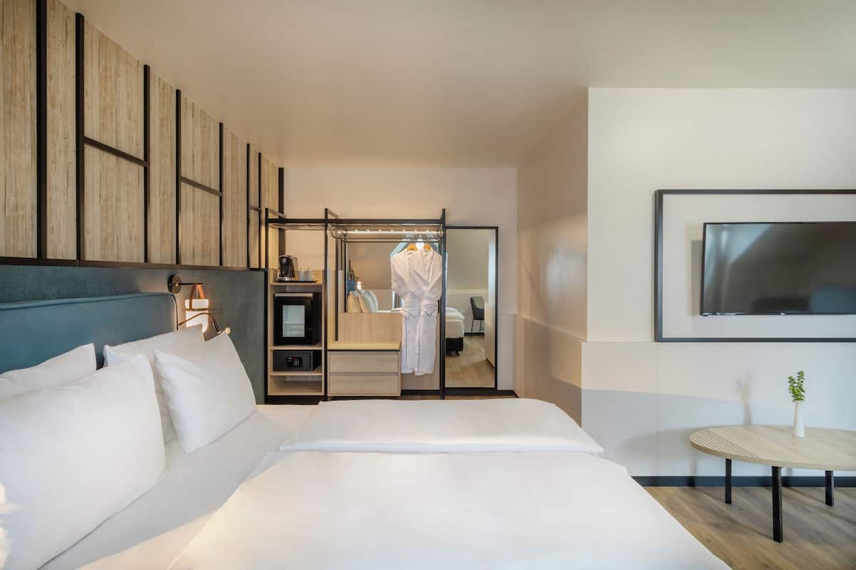 A modern hotel room with a large bed, a seating area with a table, a TV mounted on the wall, and a wardrobe with a mirror.
