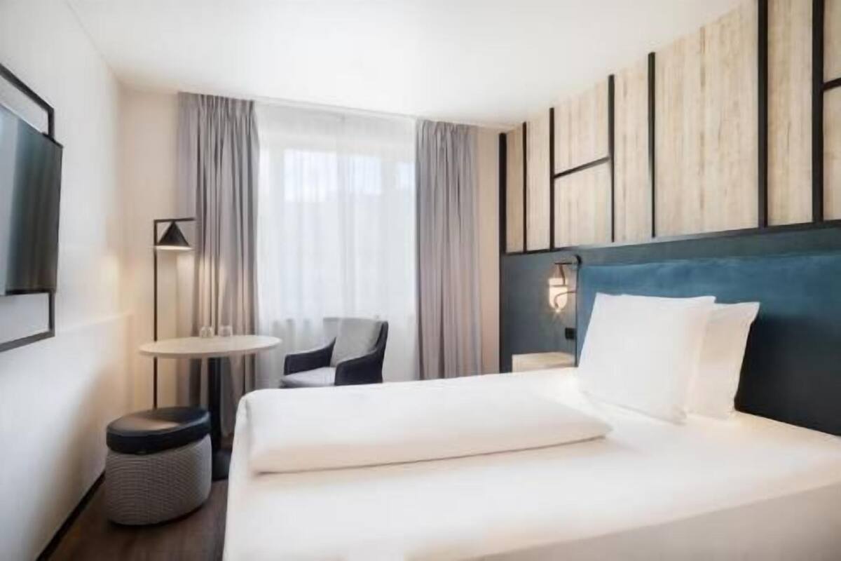 A hotel room with a large bed, a small round table, a chair, and a wall-mounted lamp.