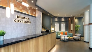 A reception area with a wooden reception desk, a sign reading "München City Center," and a seating area with a sofa and chairs. - Hotel München City Center affiliated by Meliá (Munich)