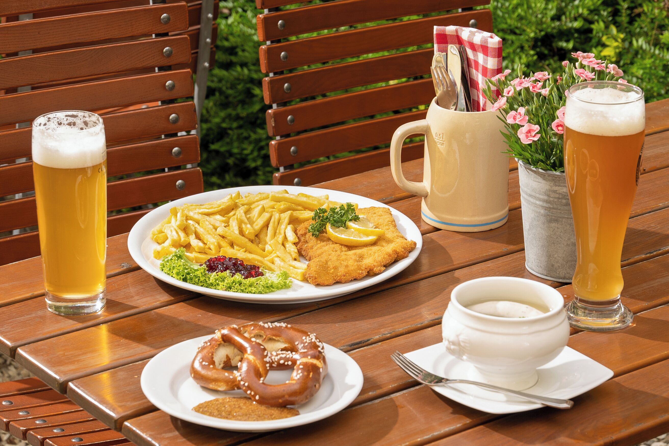 A wooden table with a meal consisting of a breaded cutlet, fries, a salad, and a pretzel. There are two glasses of beer and a mug with a spoon and fork.