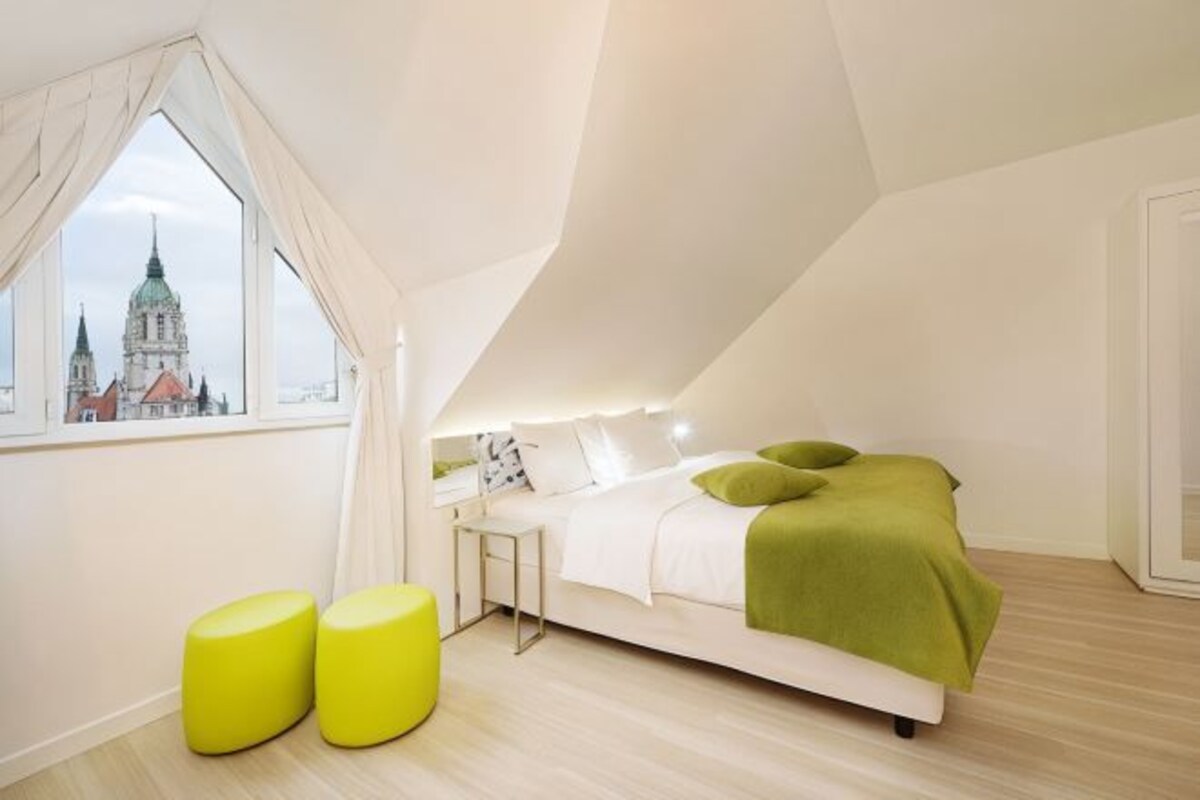 A modern bedroom with a bed, two yellow ottomans, a nightstand, and a window with a view of a cityscape.