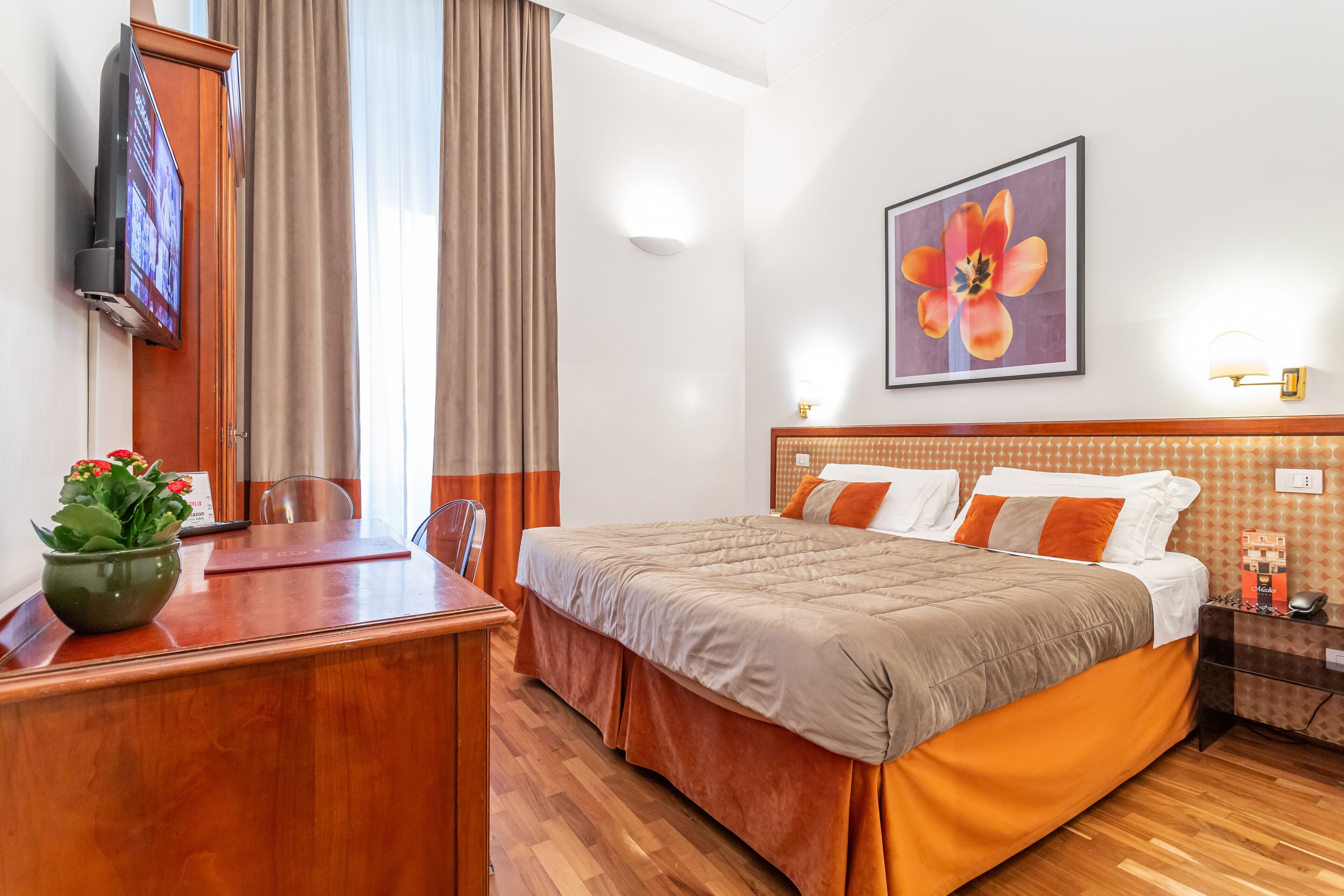 Habitación de hotel con cama, escritorio, silla, televisor, un cuadro de flores y una mesita de noche con lámpara.