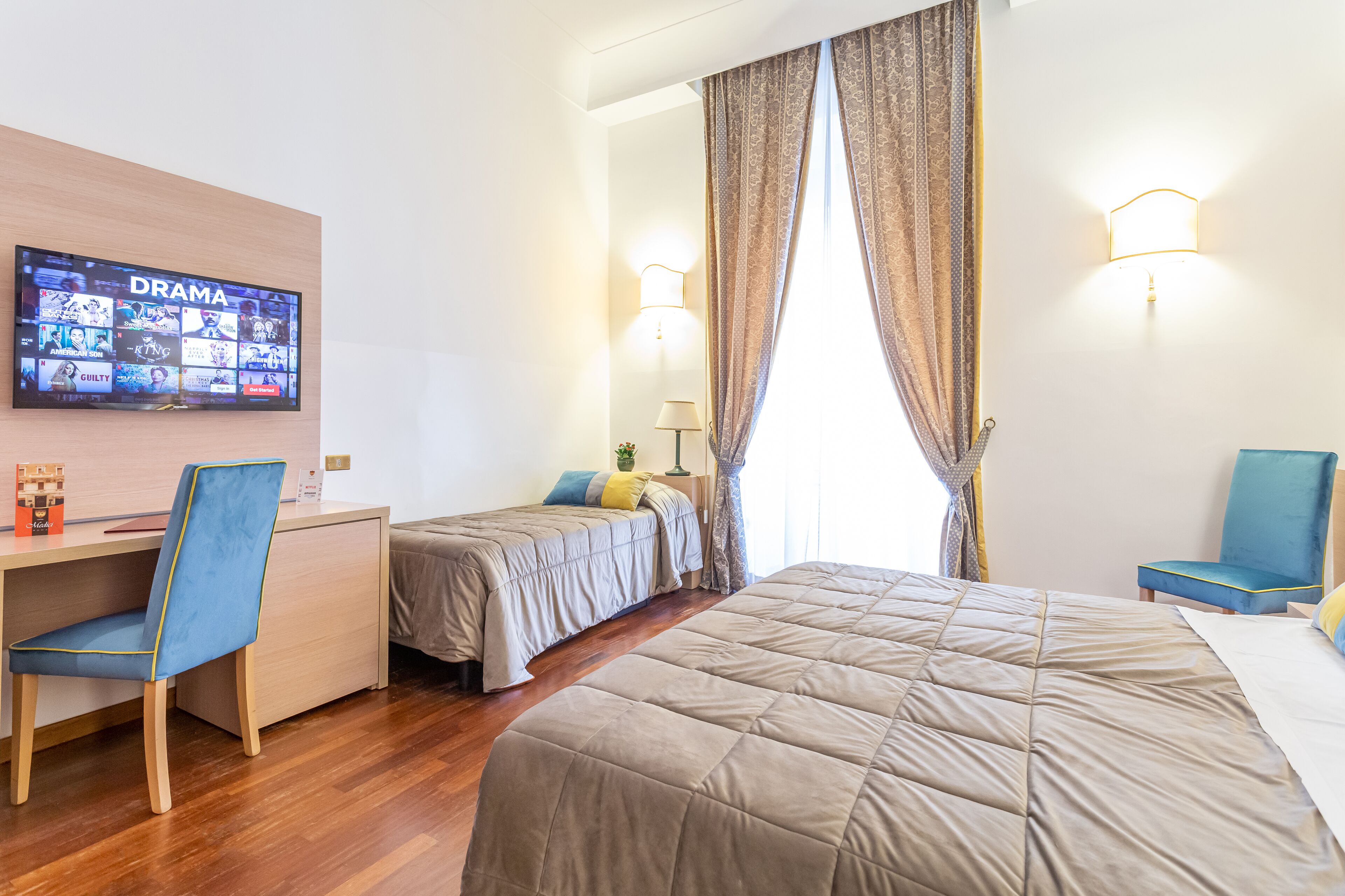 Una habitación de hotel con una cama grande, un escritorio con una silla, dos camas individuales, un televisor mostrando un canal de drama y una ventana con cortinas.