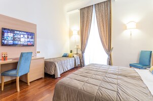 Et hotelværelse med en stor seng, et skrivebord med stol, to enkelt senge, et fjernsyn der viser en dramaserie, og et vindue med gardiner.