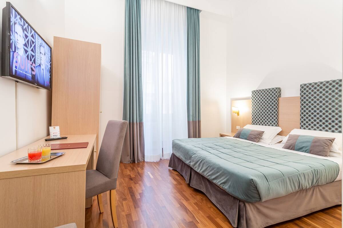 A hotel room with a bed, a desk, a TV, and a window with curtains.