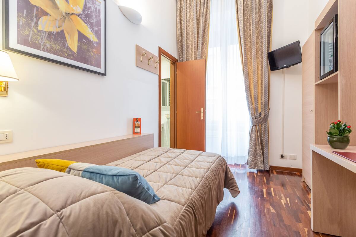 A hotel room with a bed, a TV mounted on the wall, a wooden desk, and a window with curtains.