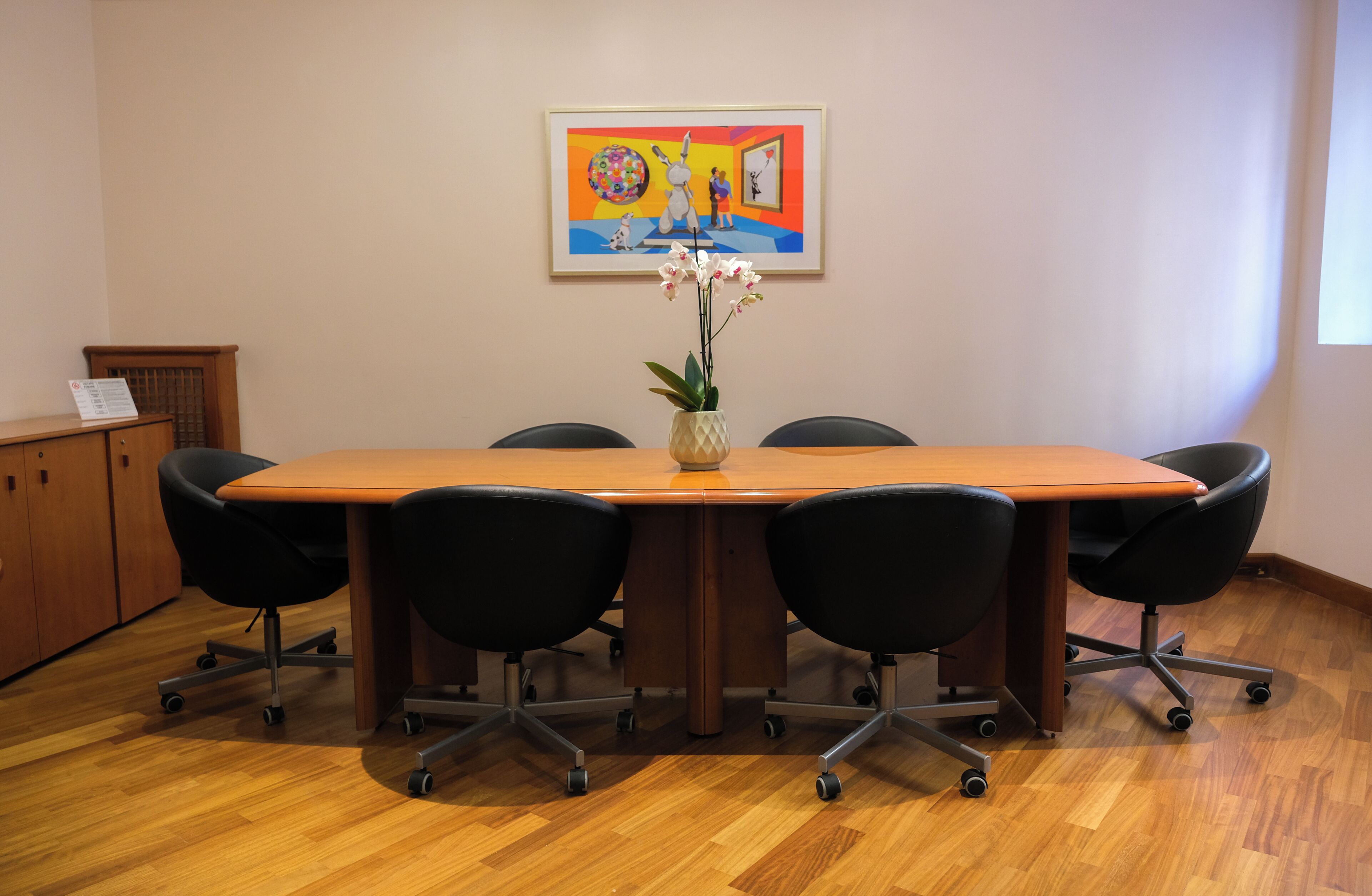 A conference room with a wooden table, black chairs, a vase with flowers, and a colorful painting on the wall.
