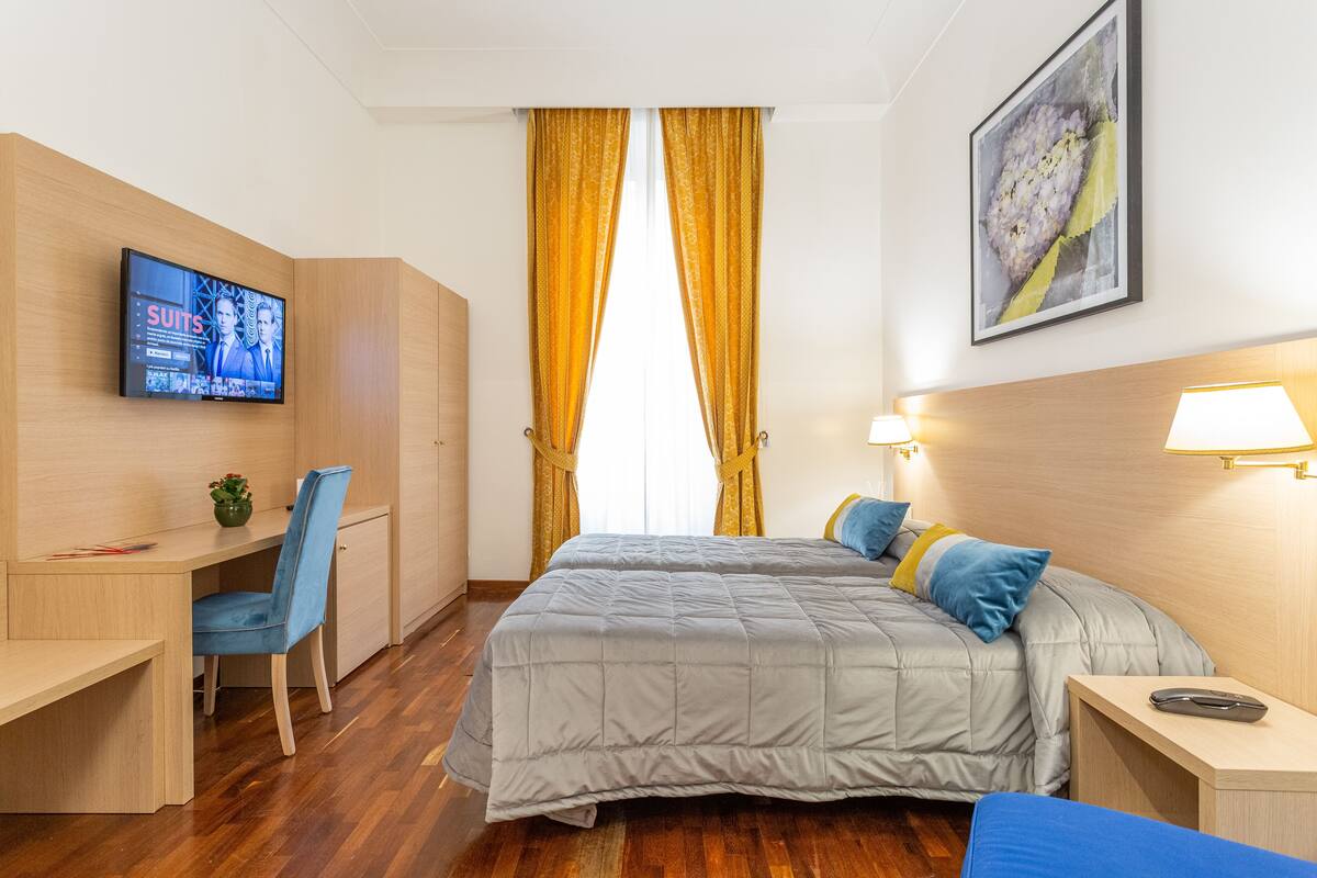 A hotel room with a bed, a desk, a chair, a TV, and a painting on the wall.