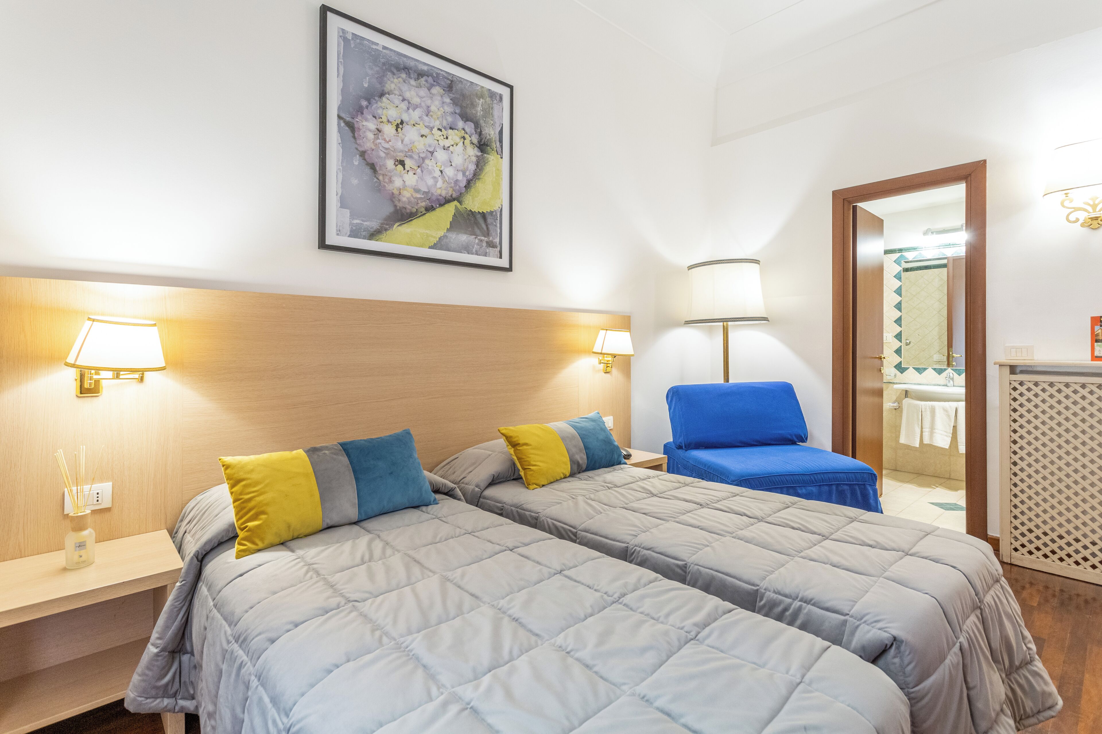 Habitació d'hotel amb dos llits, una cadira blava, una obra d'art emmarcada i un bany visible a través d'una porta oberta.