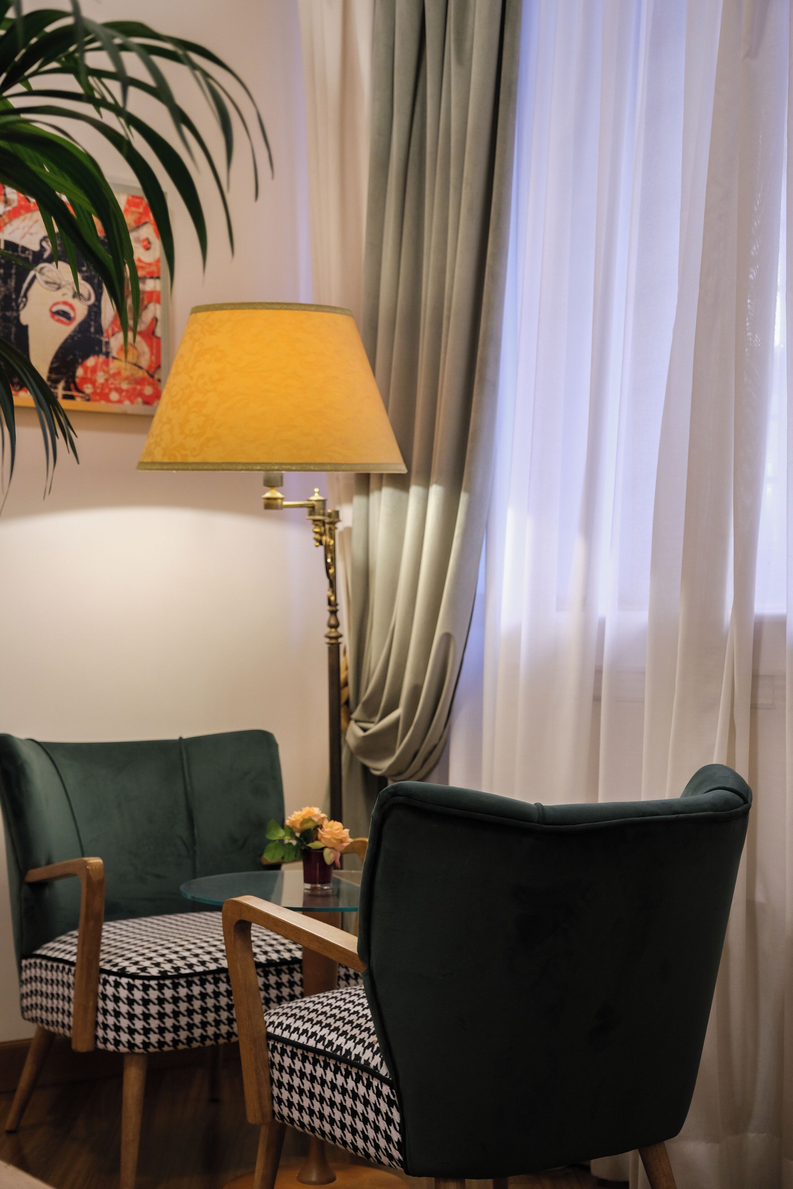 A room with a green armchair, a patterned chair, a lamp, a plant, and a painting on the wall.